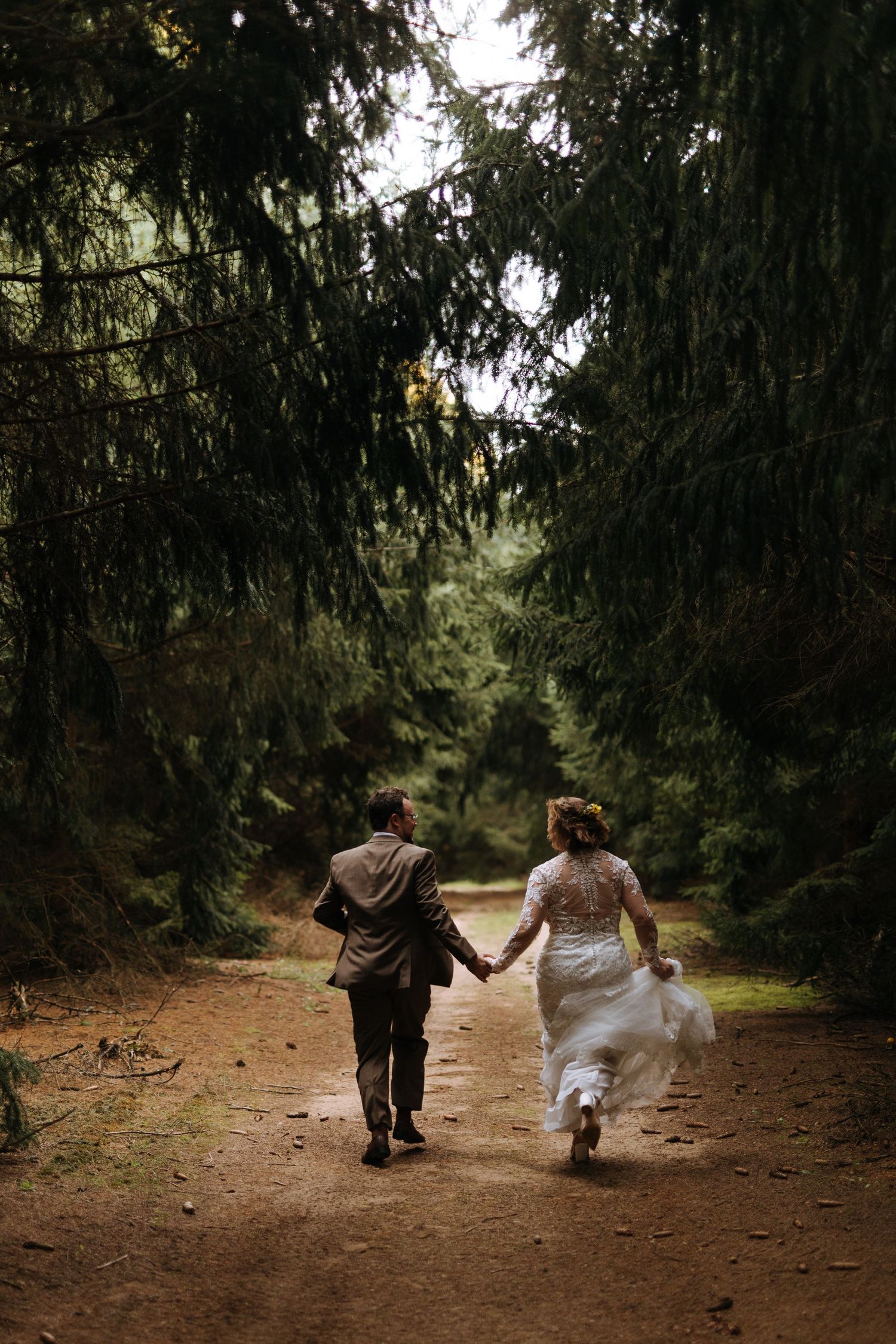 Couple walks hand in hand down forest path surrounded by tall evergreen trees.