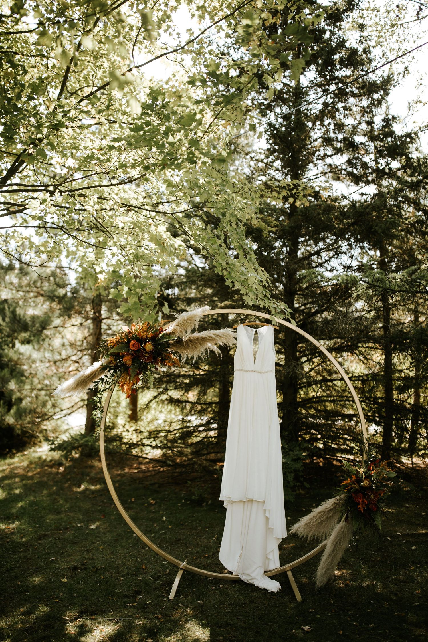 Wedding dress hanging on circular arch with floral accents in outdoor forest setting.