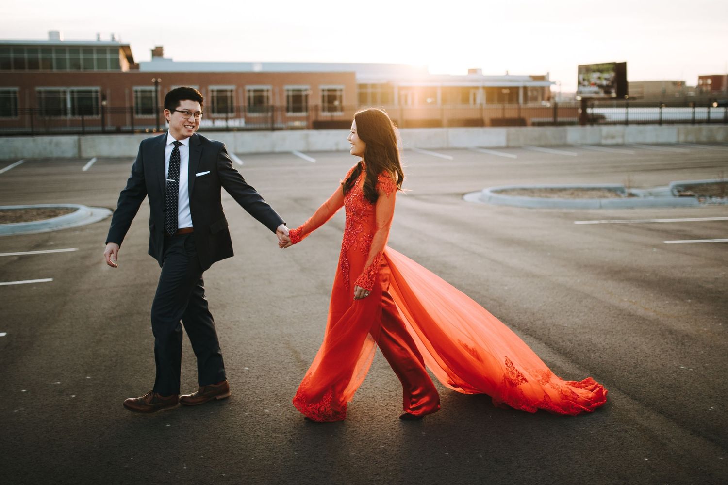 A couple in formal attire walks through an urban parking lot at sunset with a flowing red dress trailing behind them.