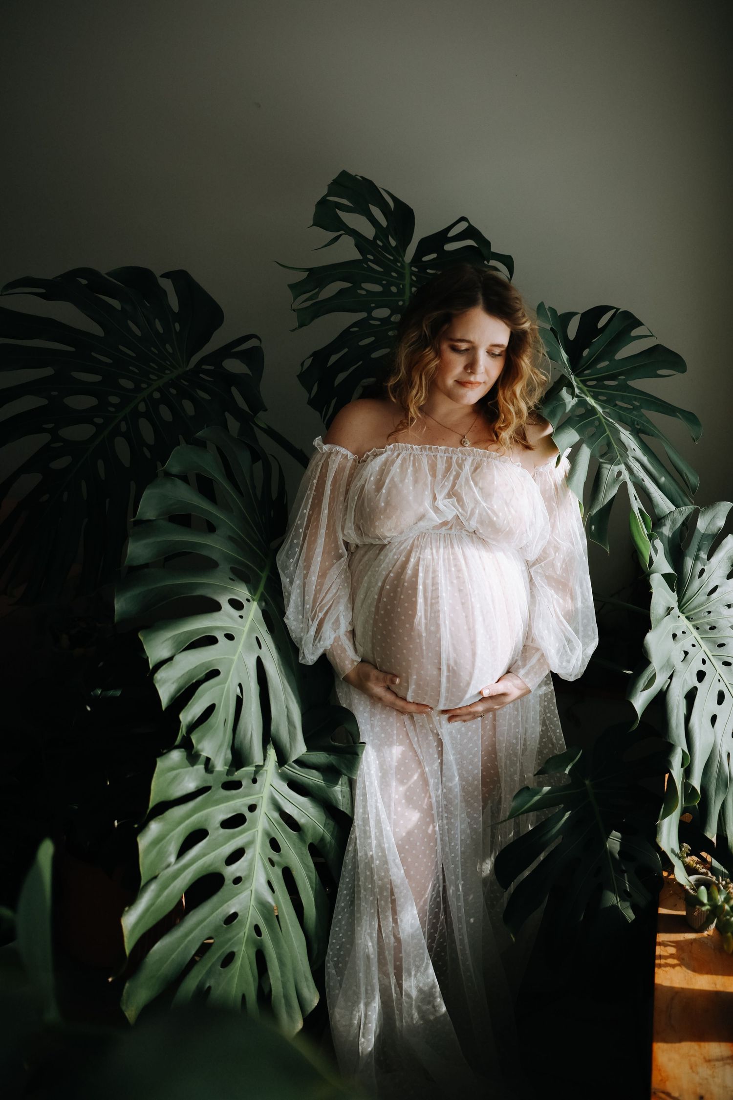 Maternity portrait with large monstera leaves against dark wall showing flowing white dress.