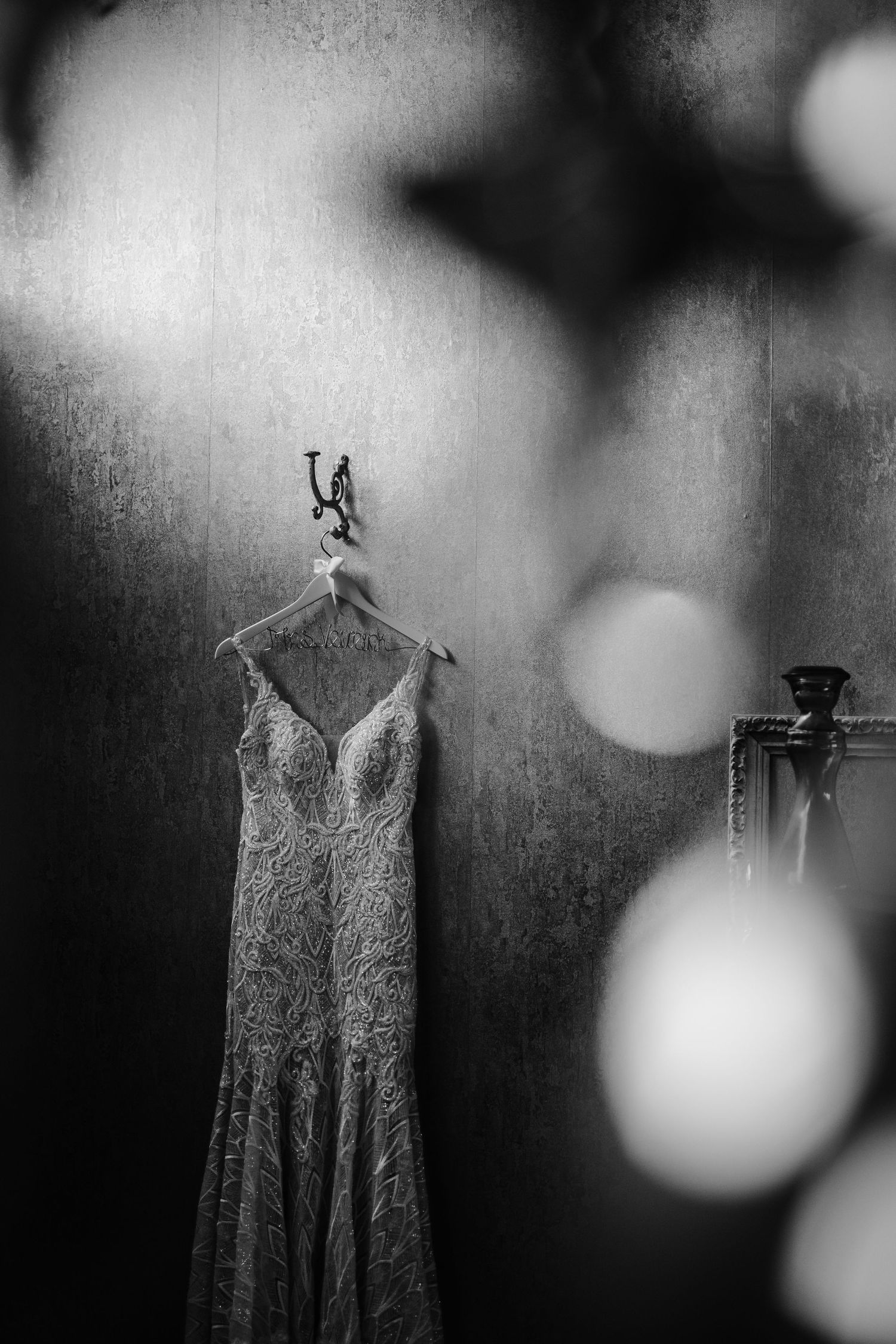 A black and white photo of a lace wedding gown hanging against a dreamy bokeh background.