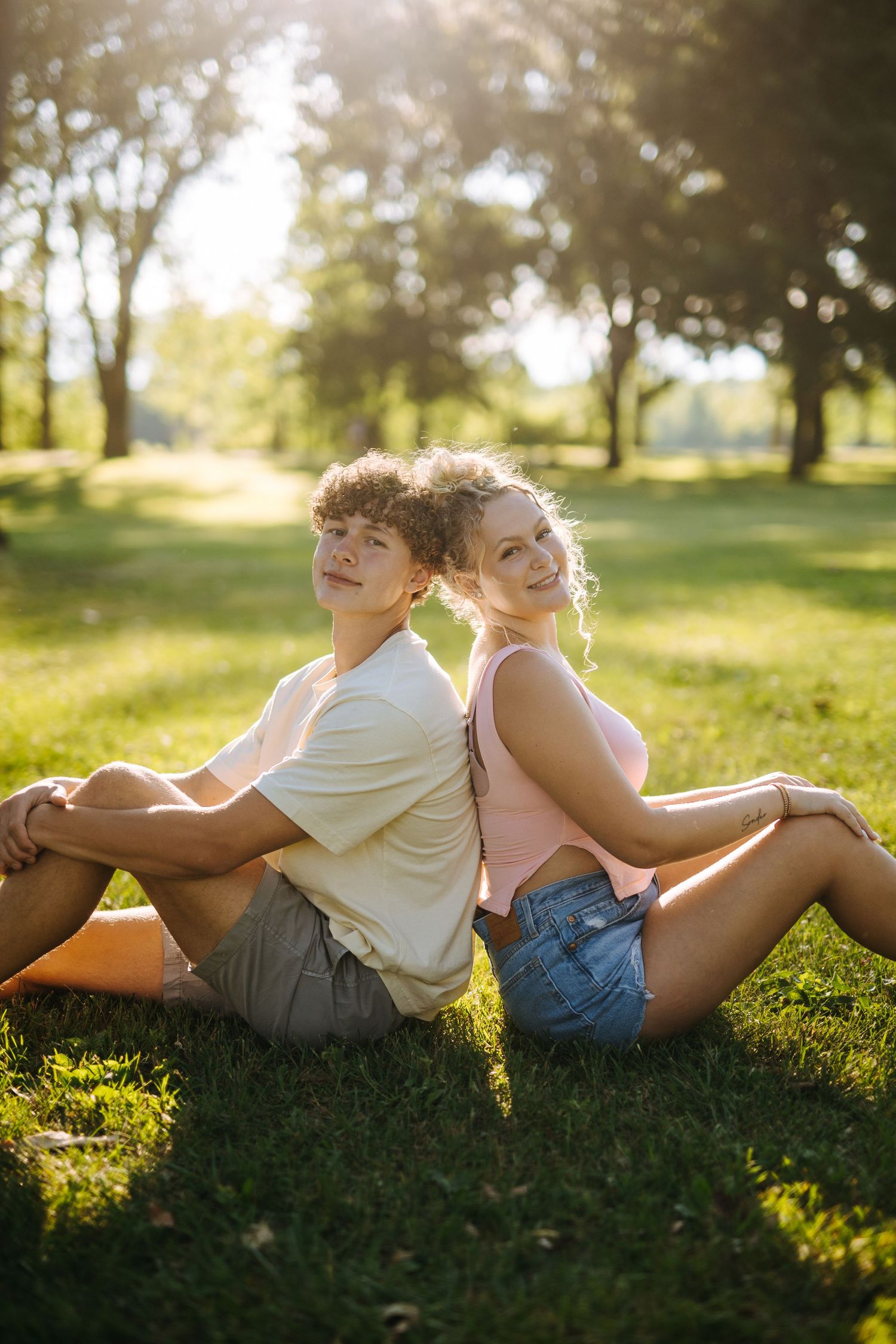 Two people laying back-to-back on green grass in a sunny park.