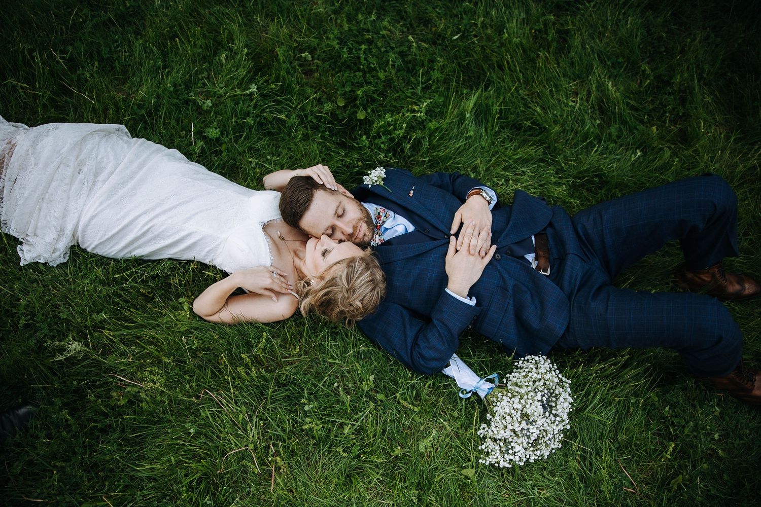 Newlyweds lay together on grass with bouquet in romantic wedding photo.
