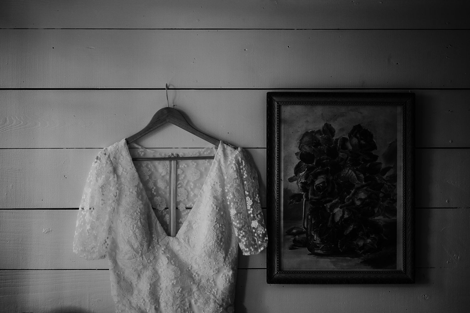 A delicate white lace wedding dress hangs elegantly against a light wooden wall with a framed artwork.