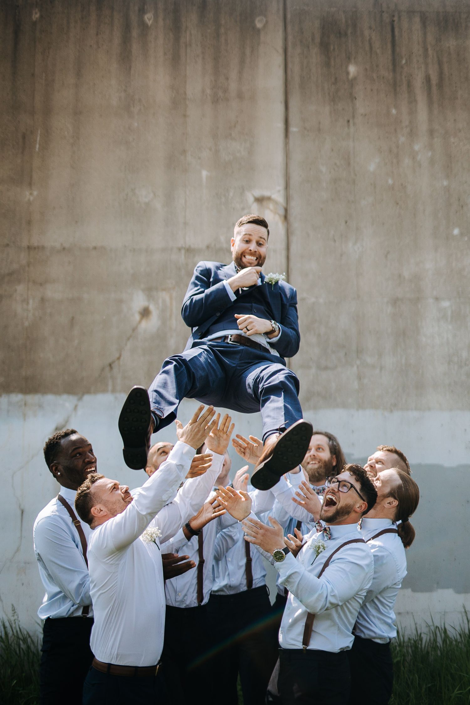 Groomsmen toss groom in air during fun wedding photo sequence against concrete wall.