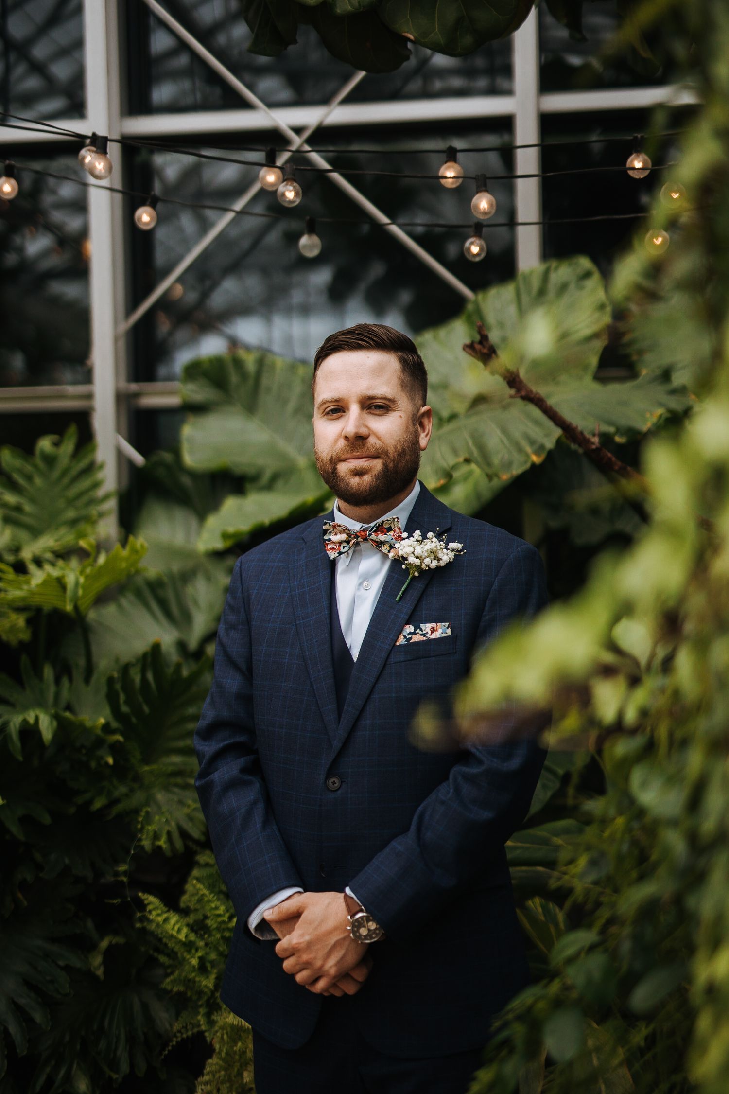 Formal wedding attire with navy suit and bow tie photographed in a greenhouse setting.
