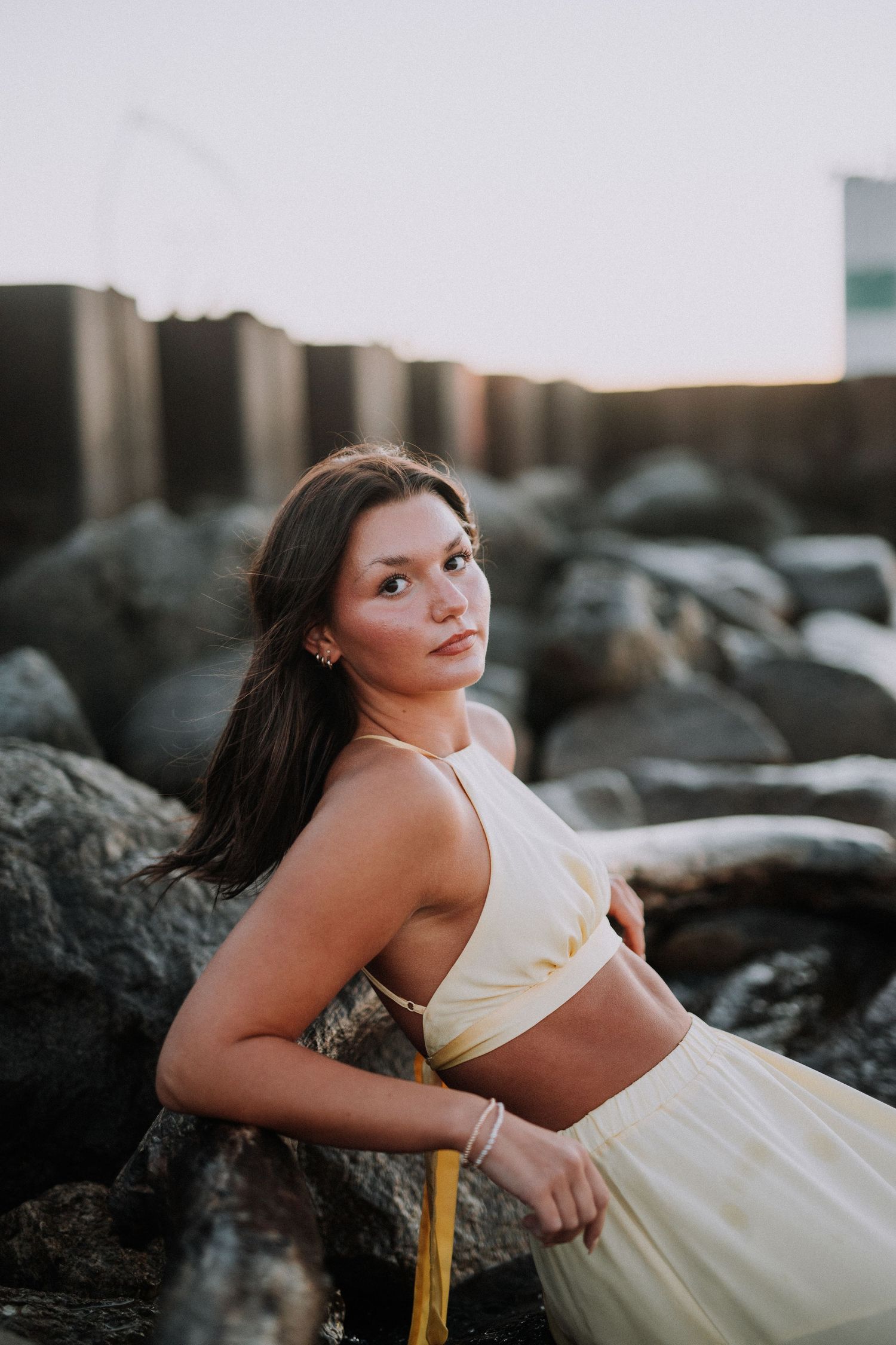 Artistic portrait on rocky beach wearing a flowing white dress at sunset.