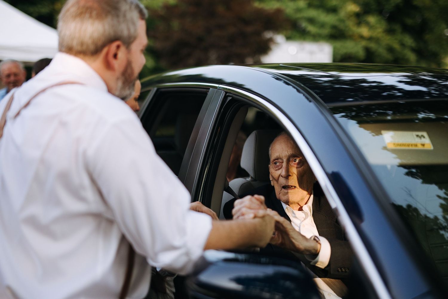 Wedding party members interact near parked vehicles during an outdoor celebration.