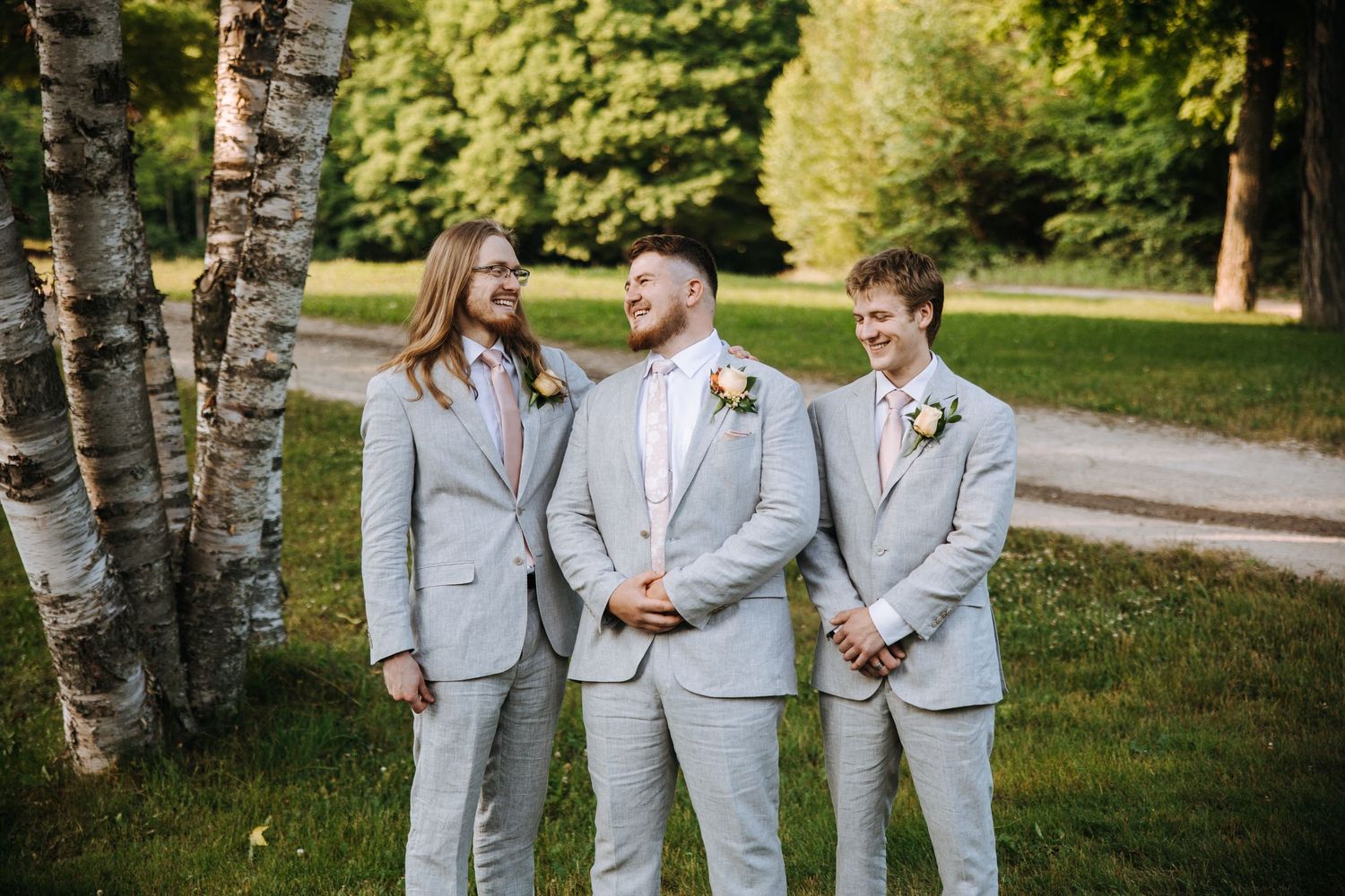 Wedding party in light gray suits poses outdoors among birch trees in a natural setting.