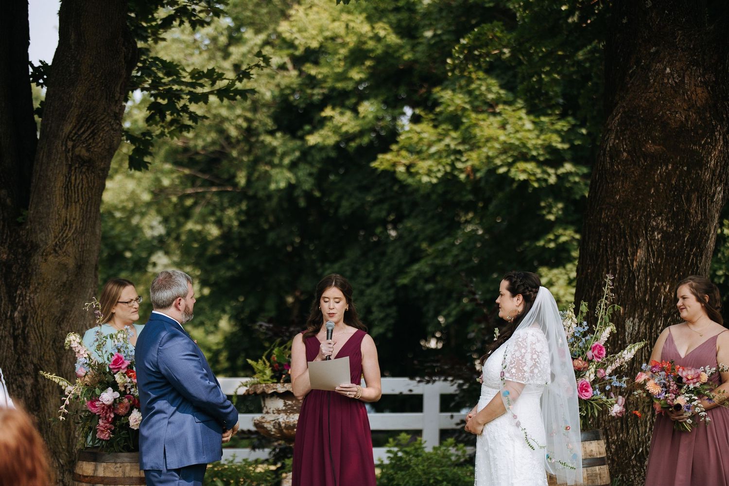 Outdoor wedding ceremony with guests gathered under mature trees on a sunny day.