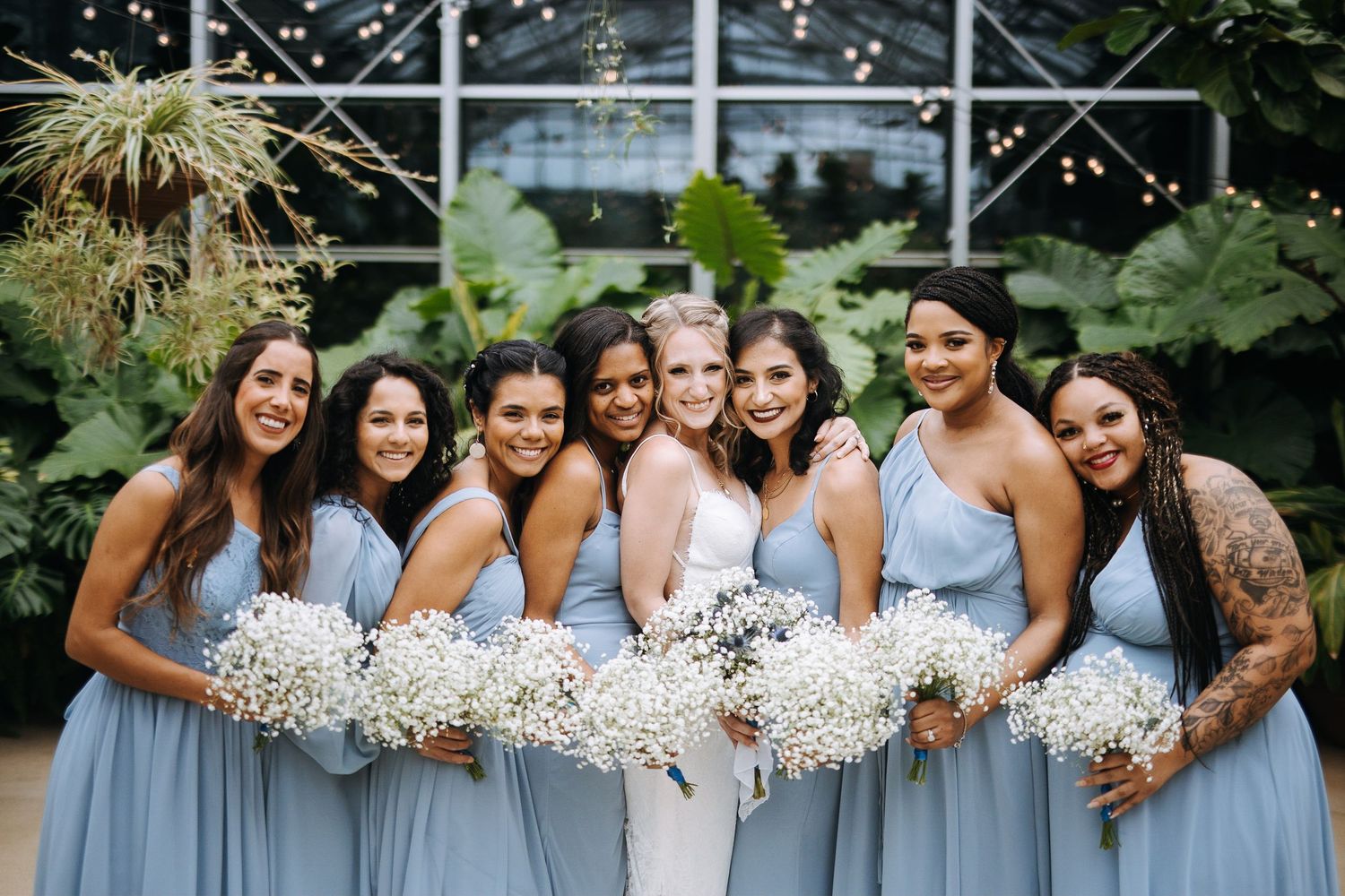 Bridesmaids in light blue dresses pose together holding white baby's breath bouquets in an outdoor garden setting.