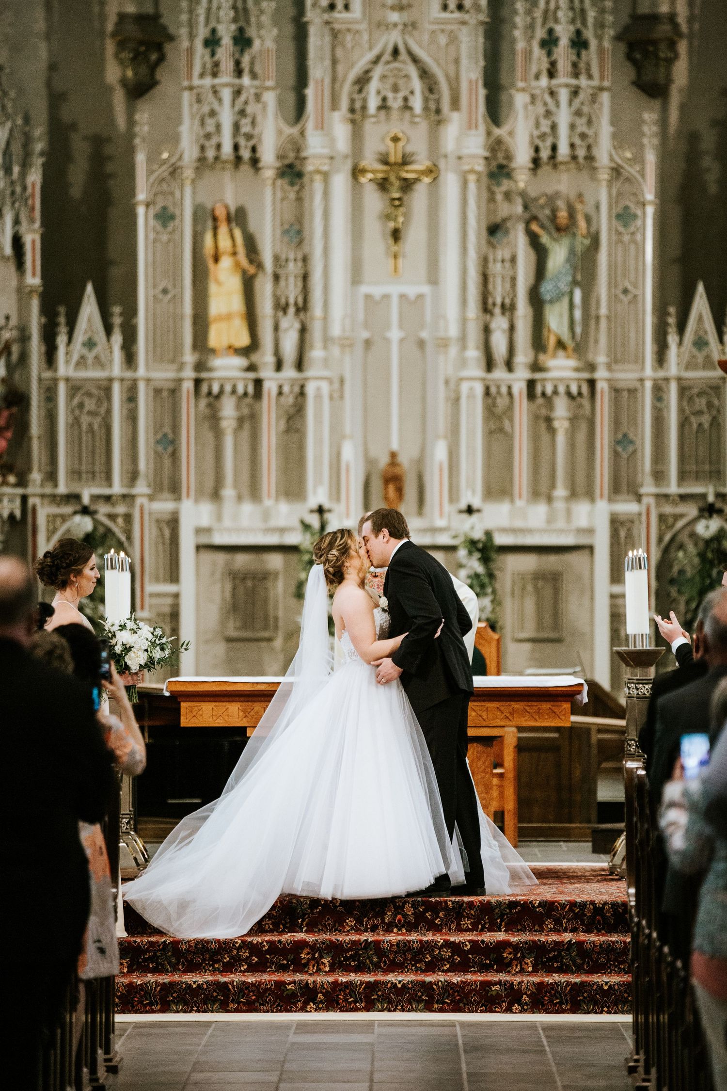 A wedding couple kneels at the church altar surrounded by ornate Gothic architecture and decorative elements.