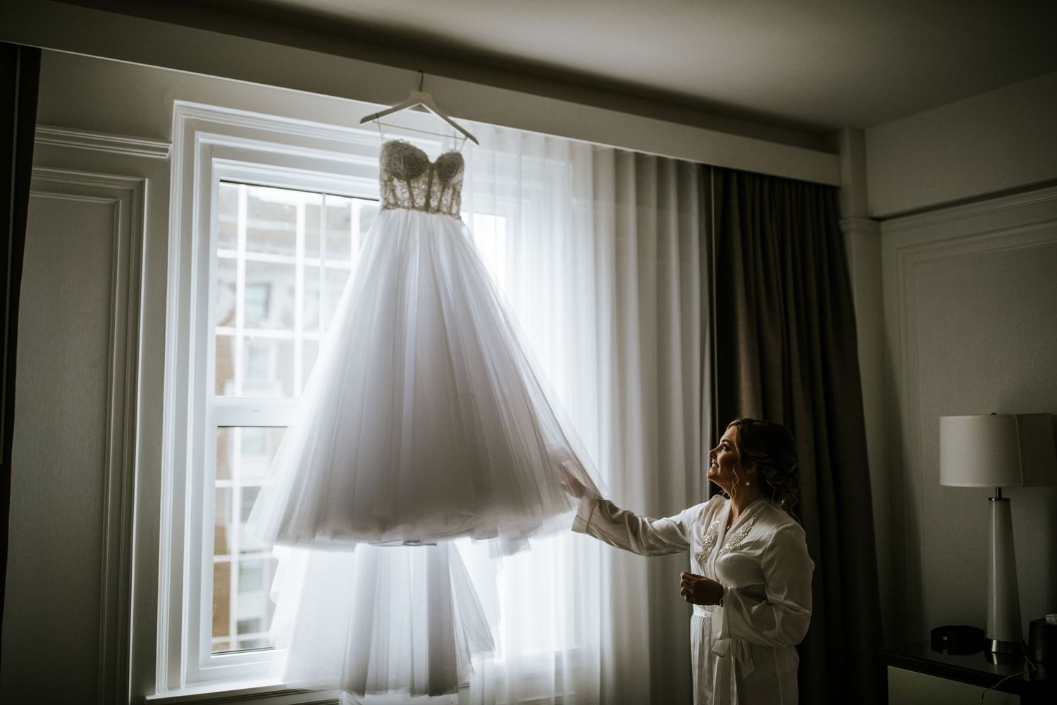 A flowing white wedding gown hangs elegantly in front of a bright window in a hotel room.