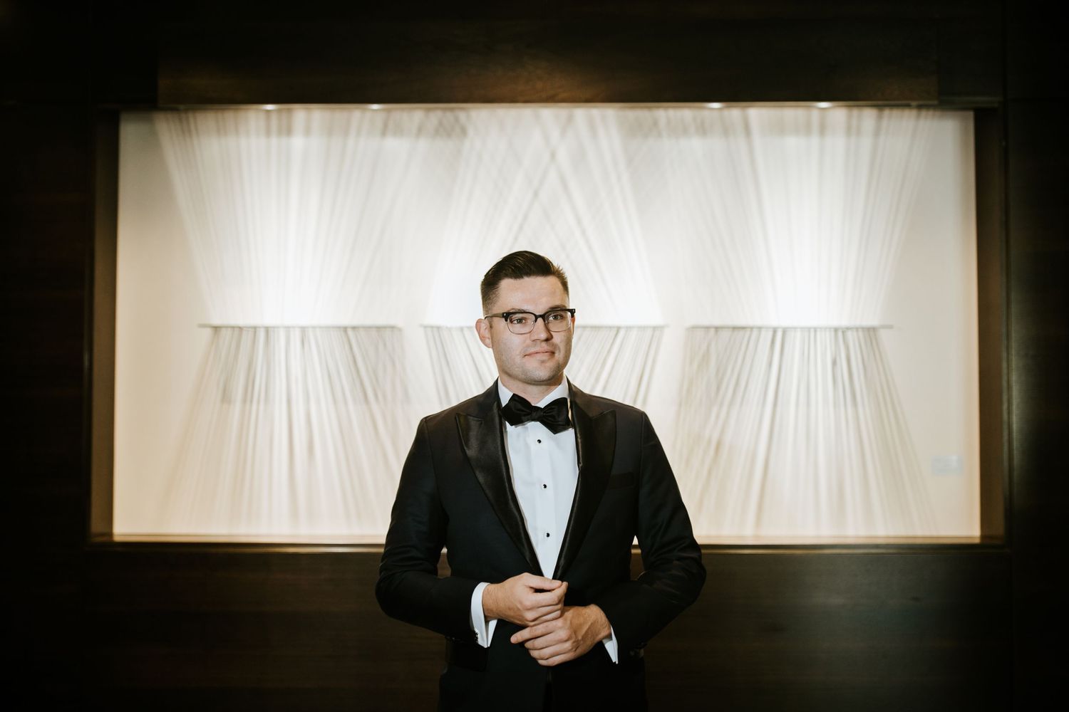 A person in a black tuxedo and bow tie stands against a white curtain backdrop with dramatic lighting.