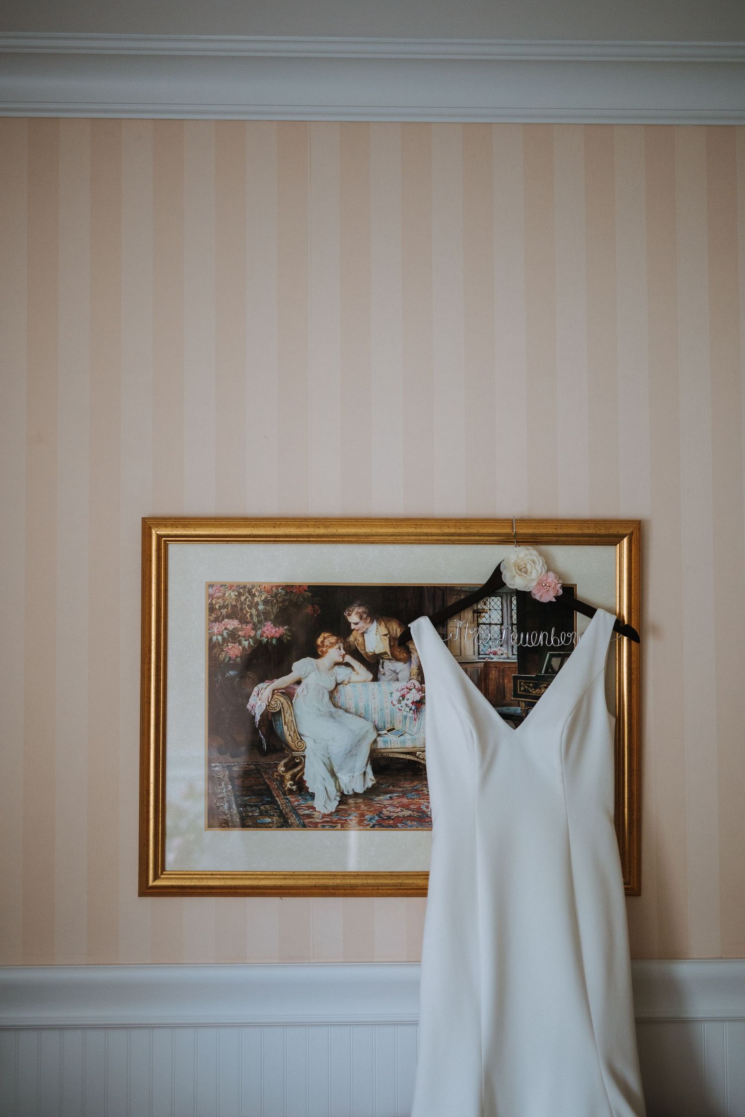 A white wedding dress hangs on display against striped wallpaper above white wainscoting.