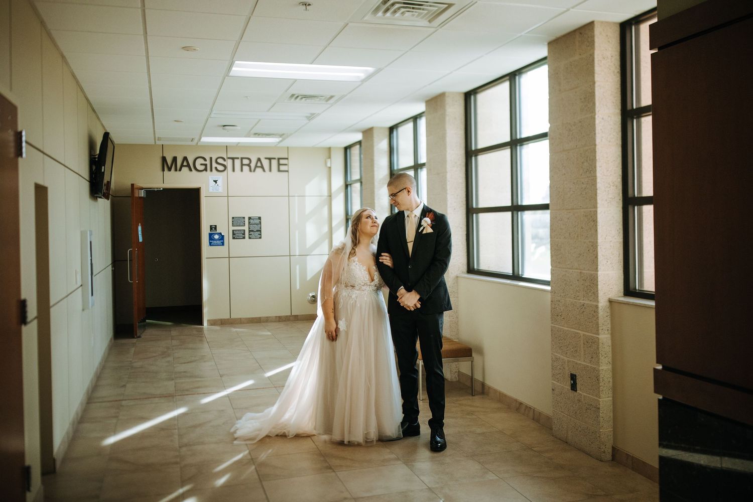 A wedding couple stands in a courthouse hallway near a magistrate office sign.