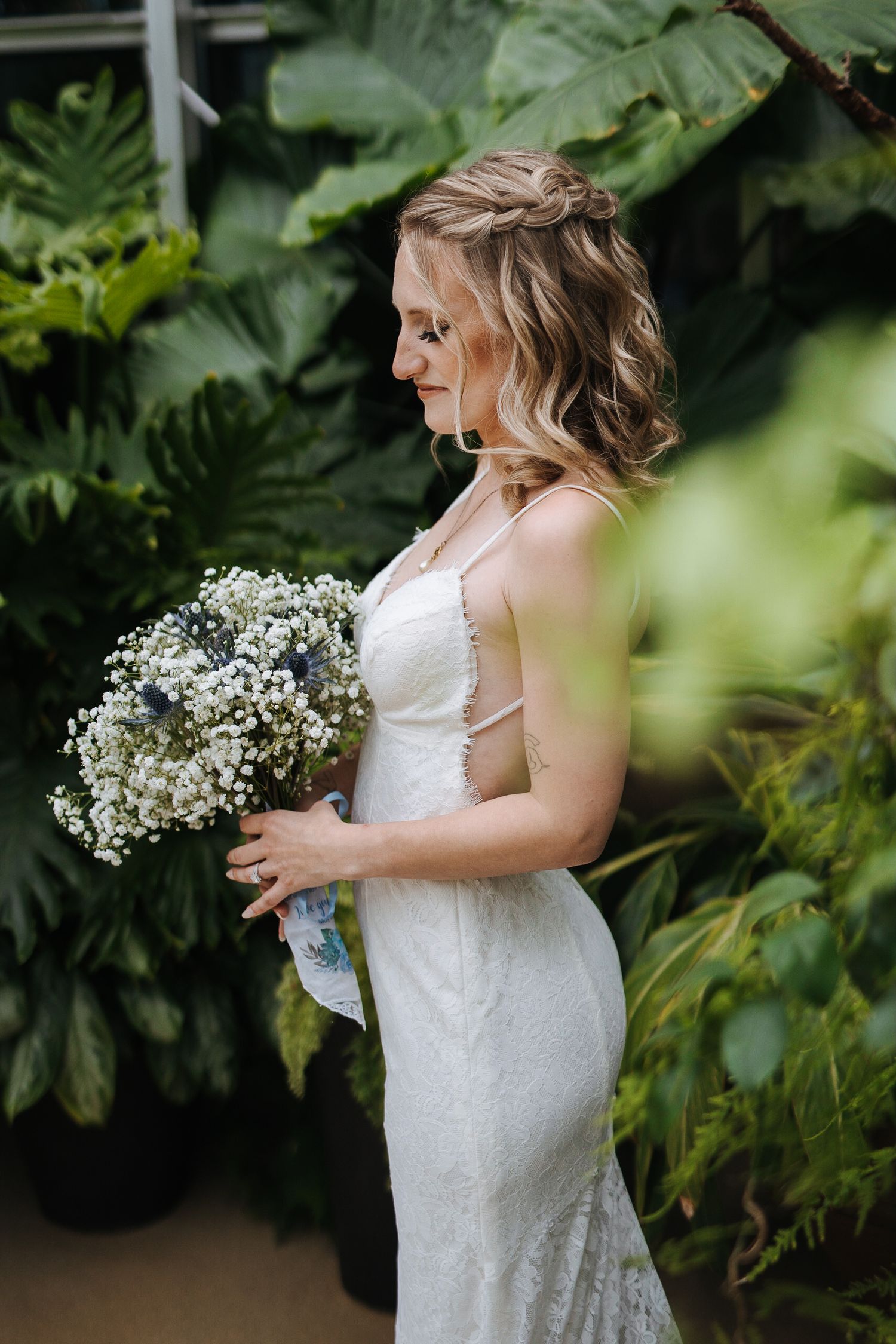 A white lace wedding dress with delicate details is photographed against lush green foliage.