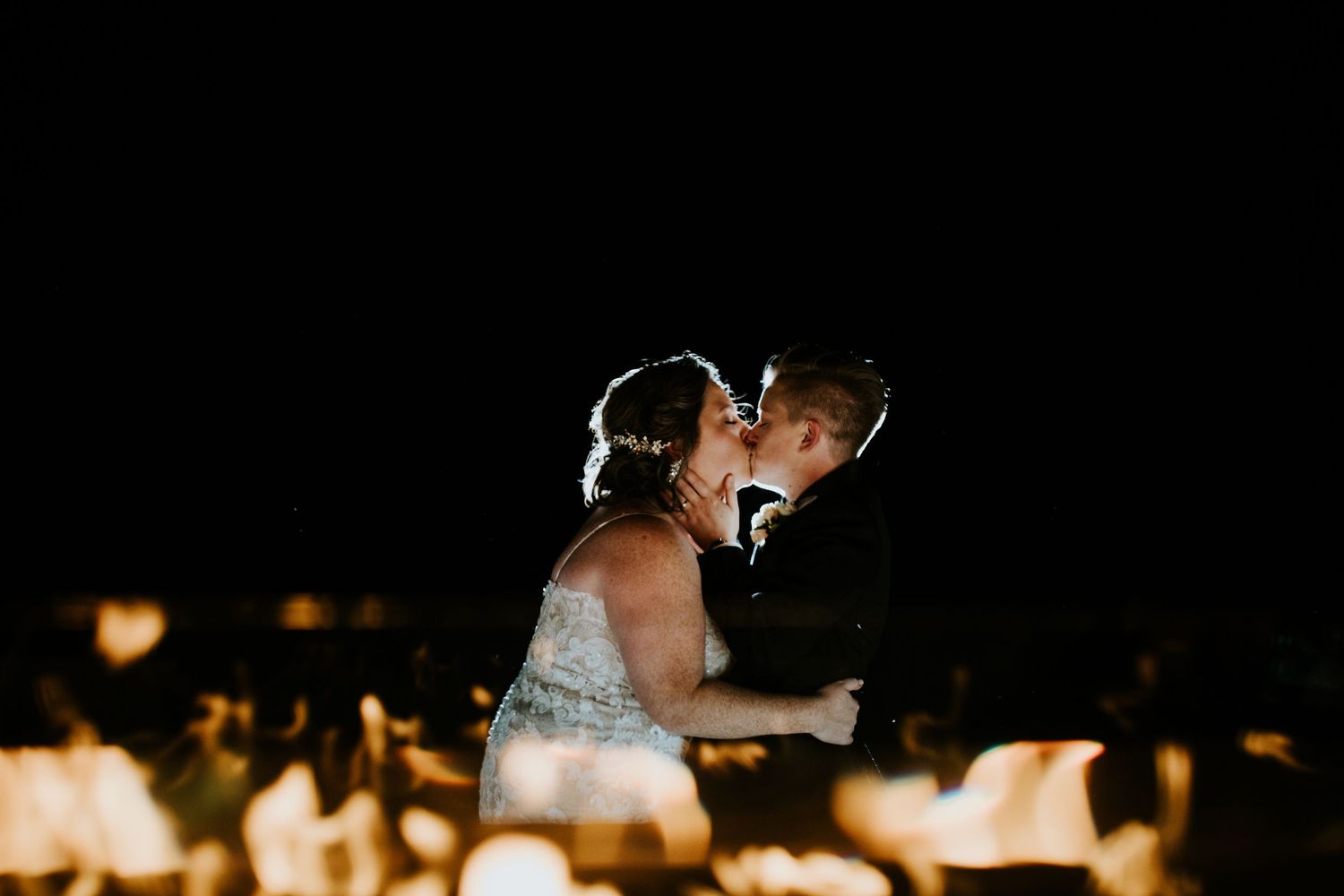 A romantic wedding couple shares a kiss in front of glowing fire pits during their evening celebration.