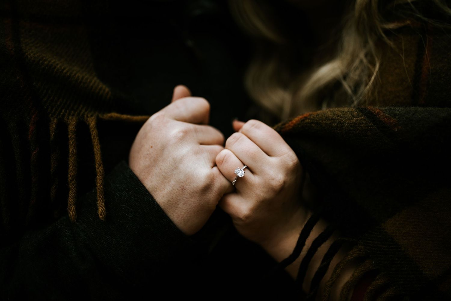 Two hands clasped together, one wearing a diamond engagement ring, against dark cozy sweaters.
