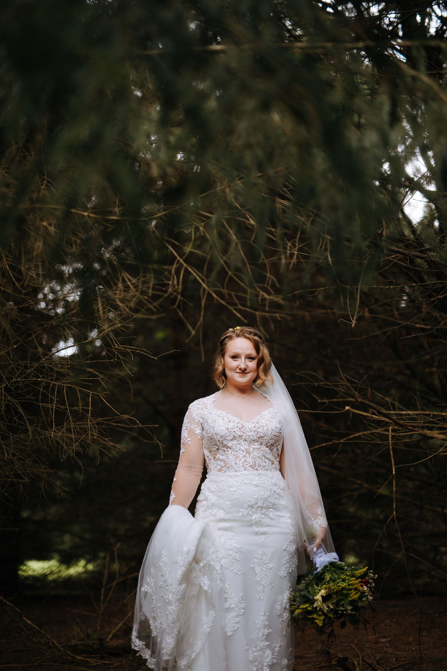 Elegant wedding dress photographed against evergreen trees in a garden setting.