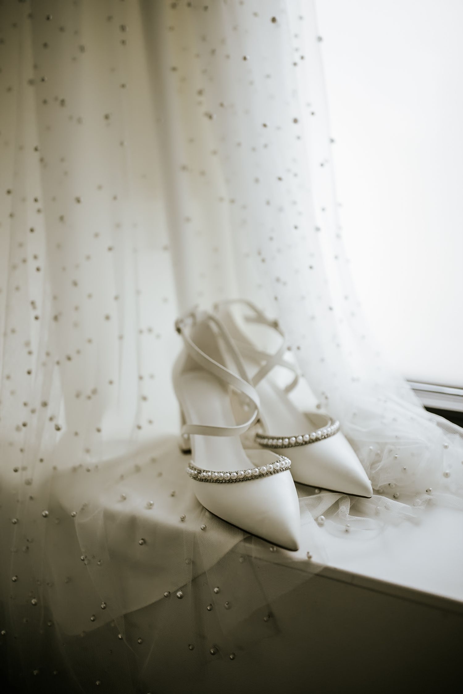 White bridal shoes with pearl details displayed on windowsill with dotted curtain.