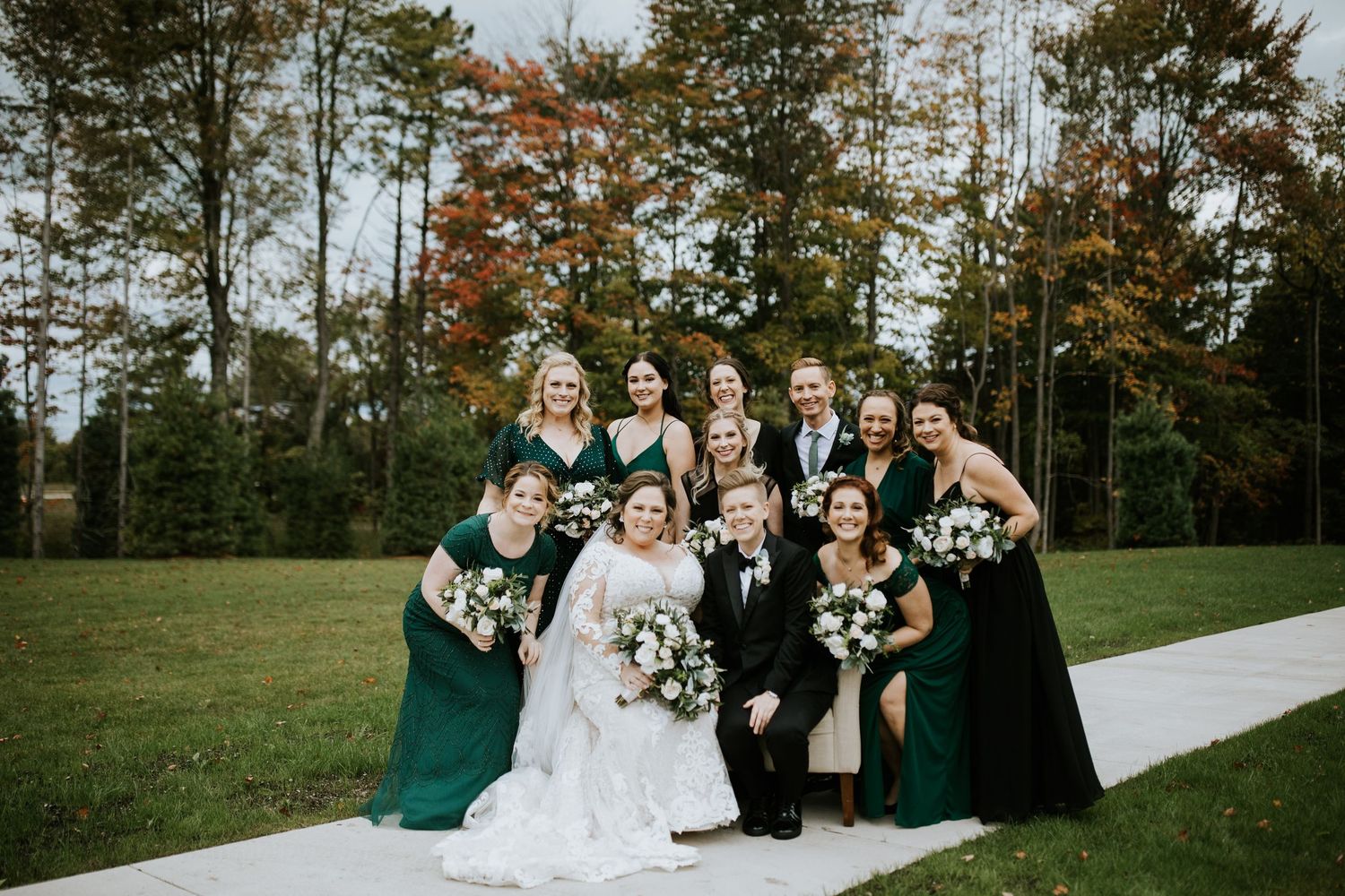 Wedding party poses together on a grassy lawn with pine trees in the background during autumn.