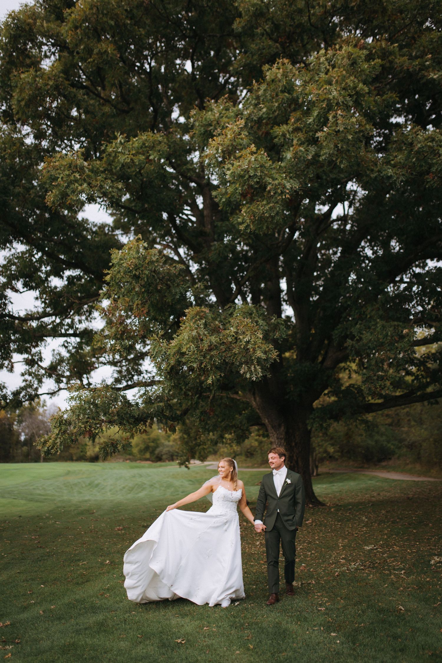 Couple walks together on grassy hill under large oak tree.