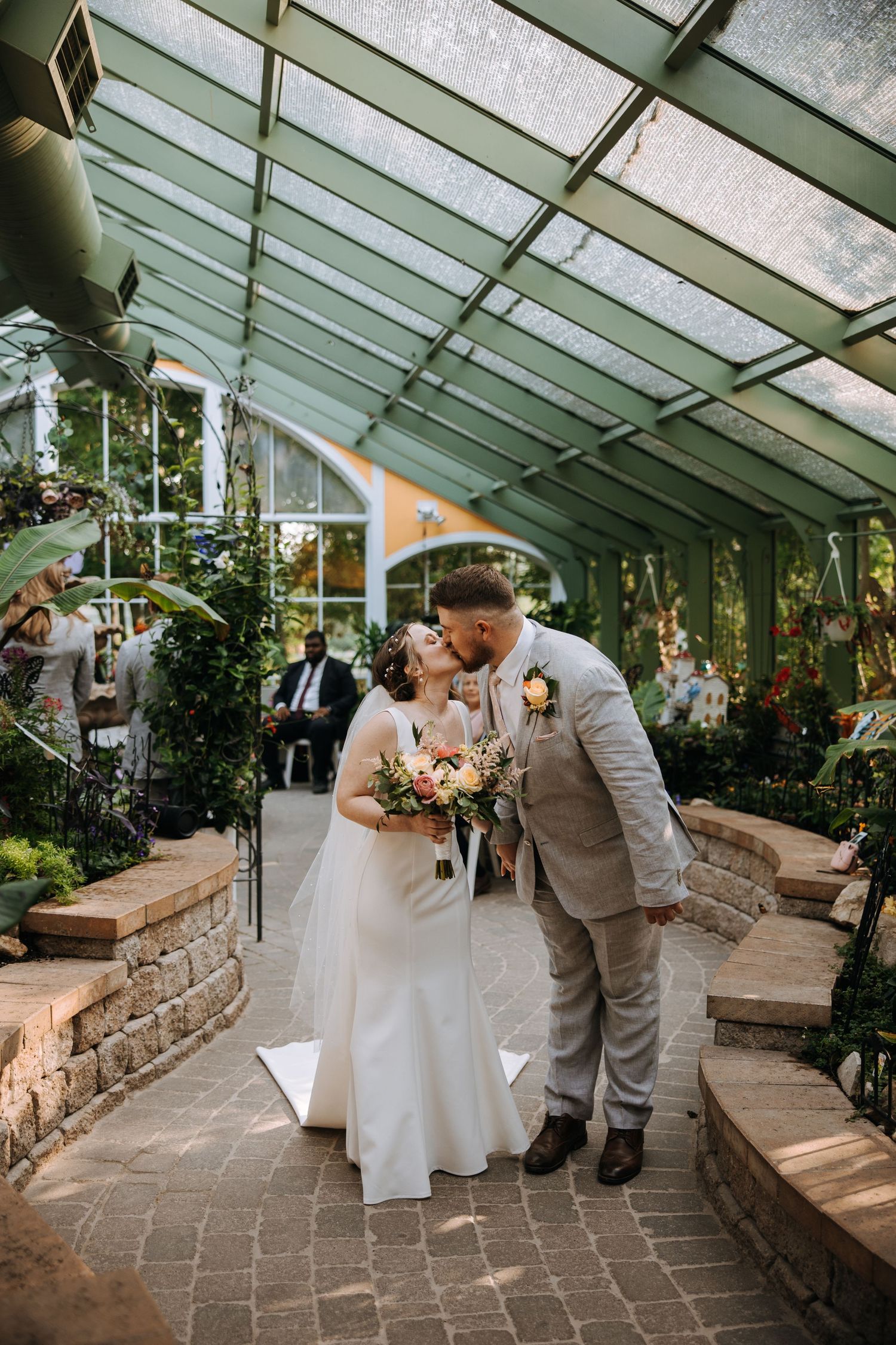 A wedding ceremony takes place in a beautiful greenhouse with stone pathways and natural lighting.