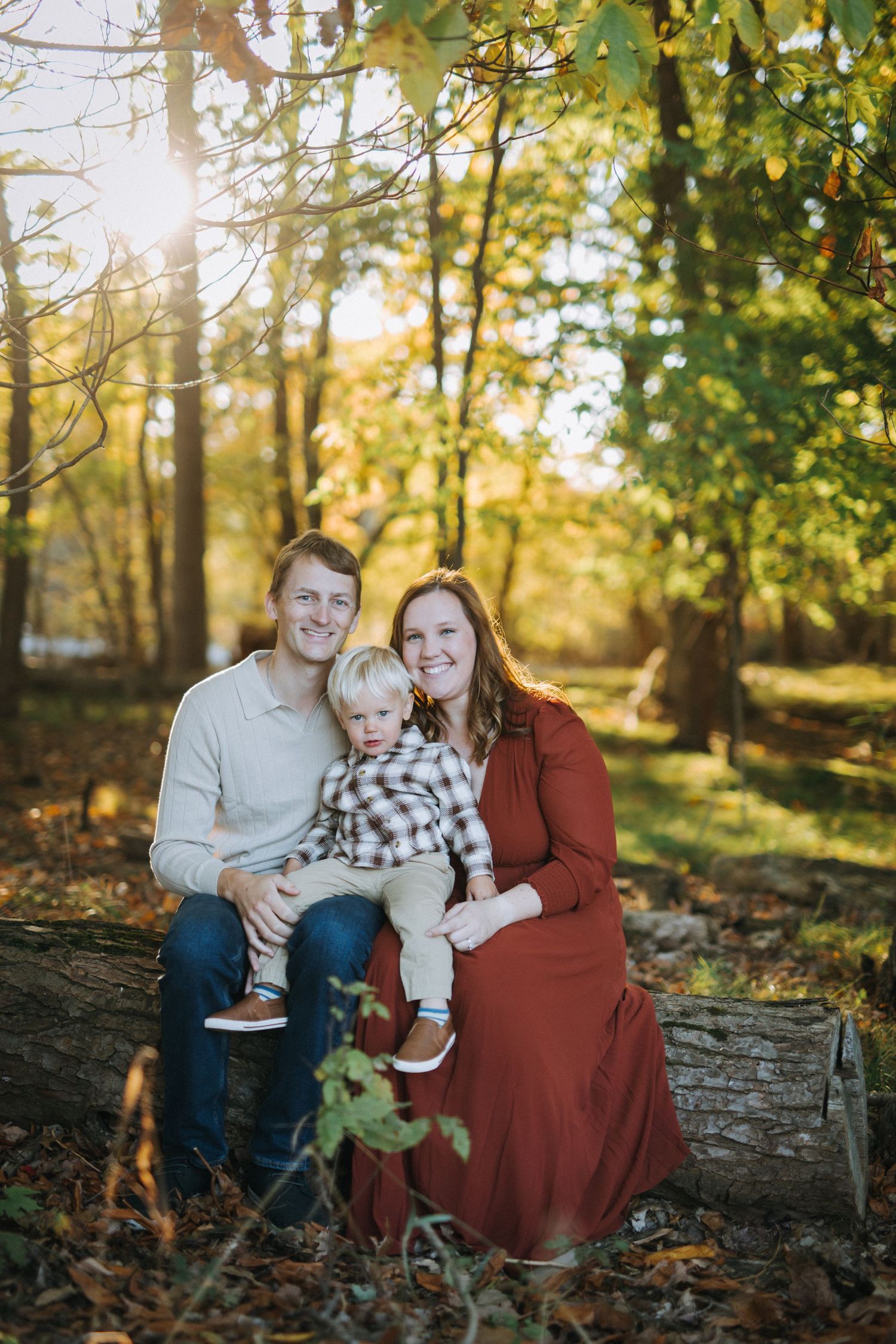 A family sits together on a fallen log in a sun-dappled autumn forest setting.