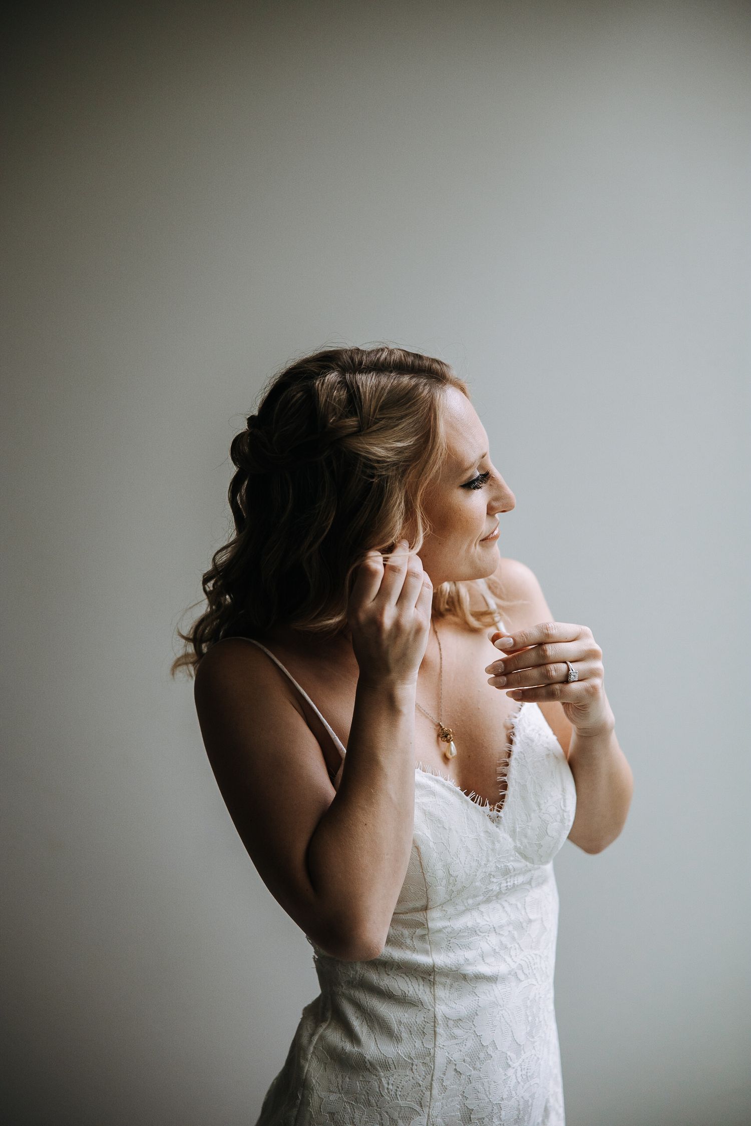 Side profile of person in white dress putting on jewelry against grey background.