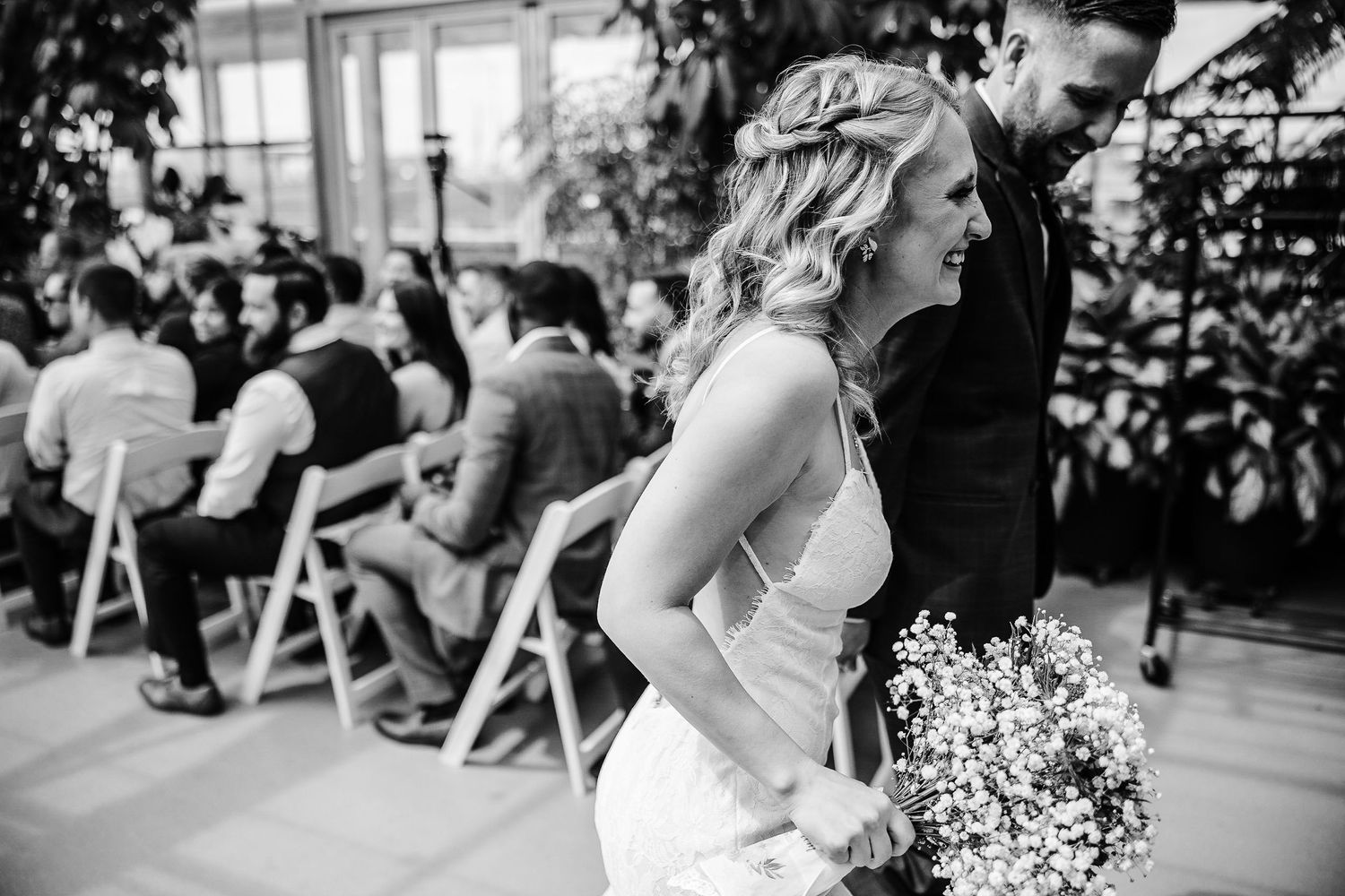 Black and white photograph of wedding ceremony guests seated in a greenhouse venue.