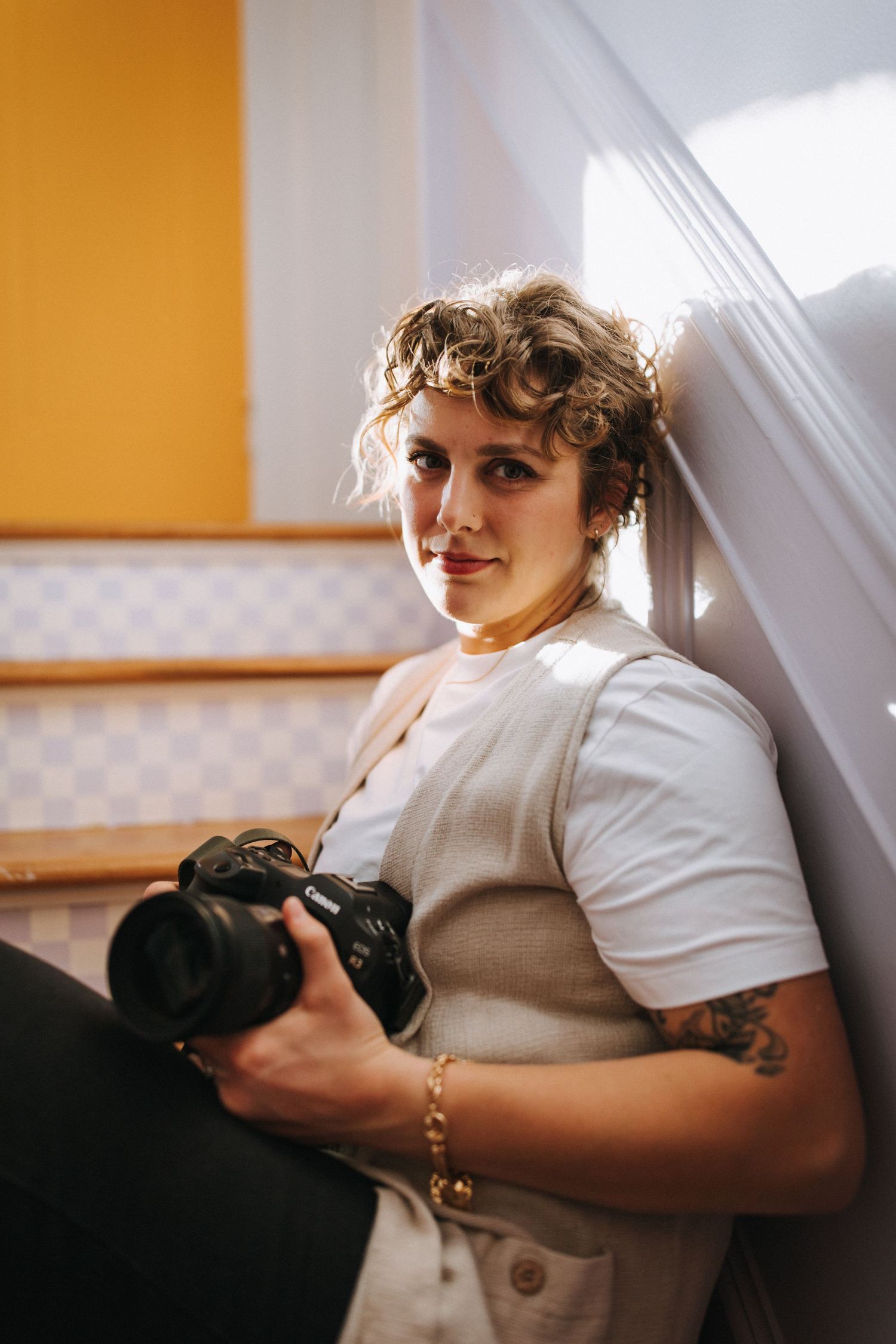A photographer with curly hair sits on stairs holding a professional camera.