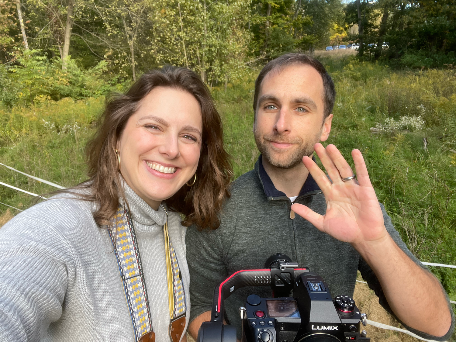 Couple taking selfies together in a sunny outdoor setting with green nature backdrop.