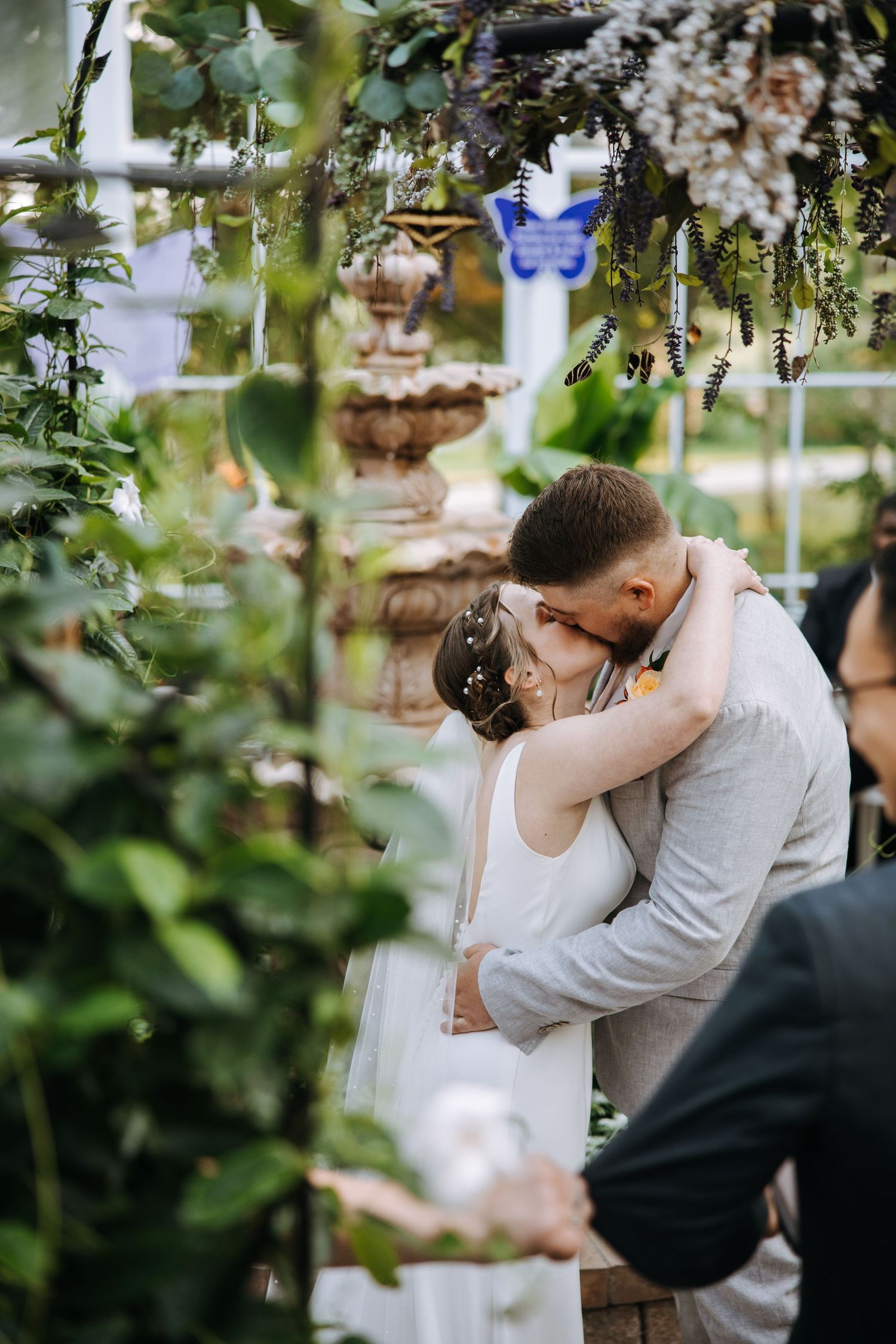 A romantic wedding embrace photographed through garden greenery with a fountain in the background.