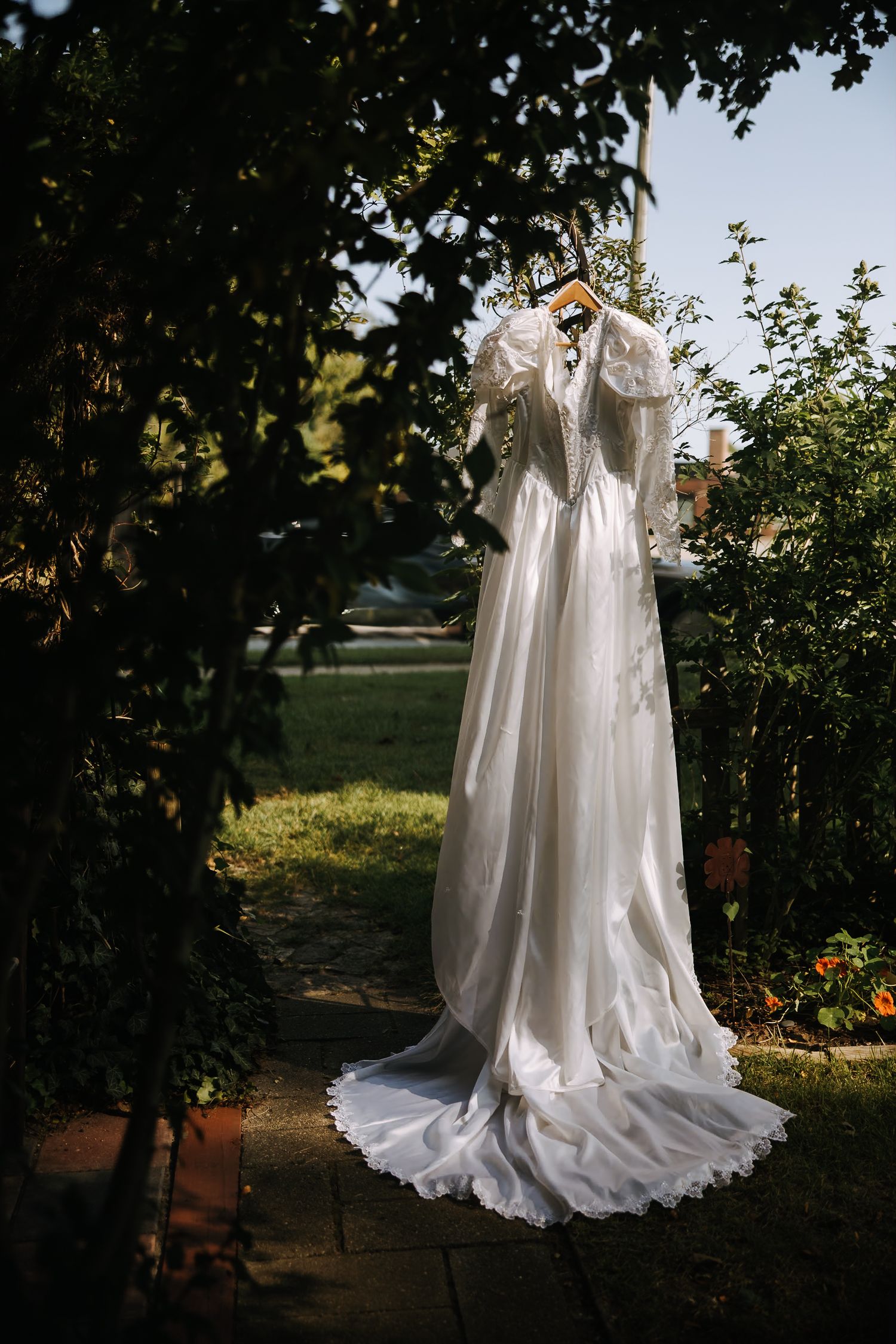 A white wedding dress hangs elegantly among green foliage in natural outdoor lighting.