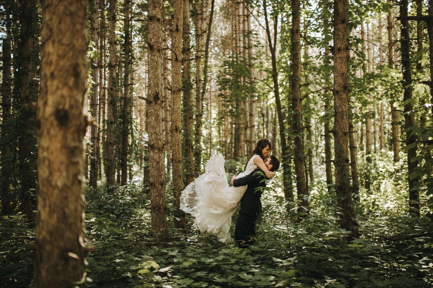 A couple in wedding attire embraces in a sunlit forest setting during their outdoor wedding ceremony.