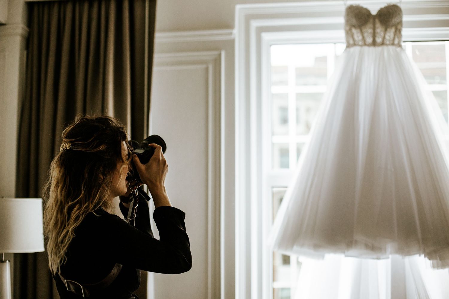 White wedding dress hanging by window with elegant curtains in natural light.