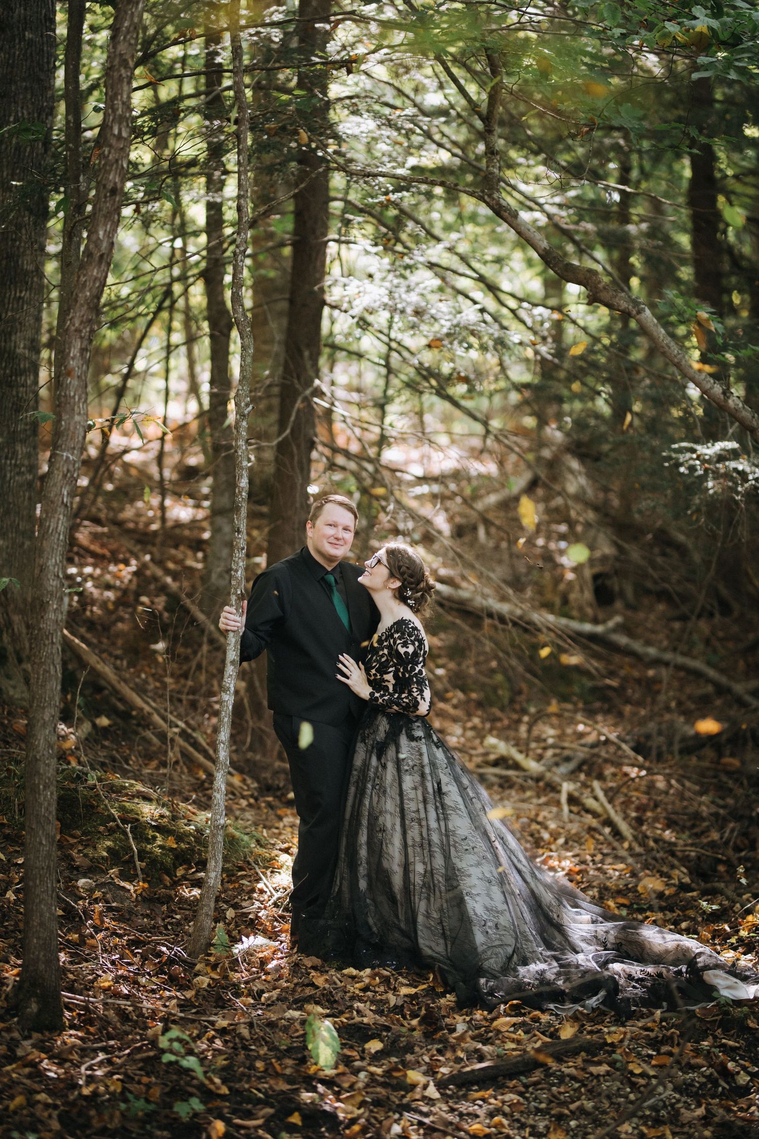 Couple in formal black attire poses romantically in wooded forest setting.