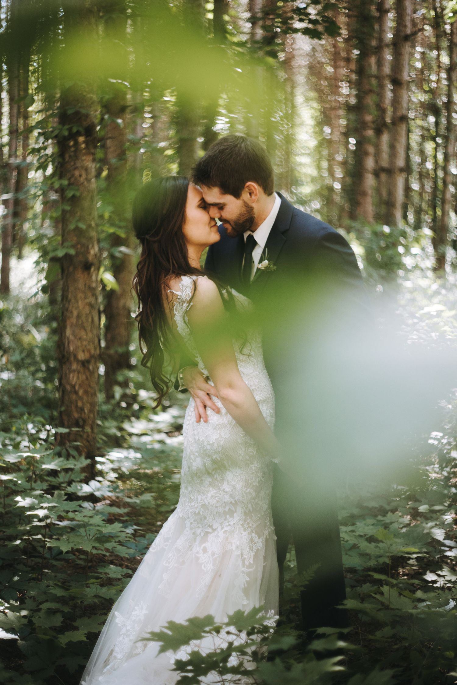 A romantic wedding couple shares intimate moments in a lush forest setting surrounded by pine trees and white wildflowers.