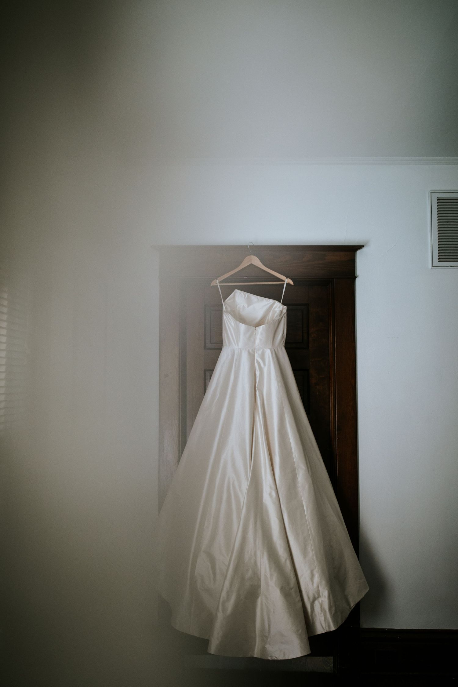 A white wedding dress hangs elegantly on a wooden hanger against a light-colored wall in a dimly lit room.