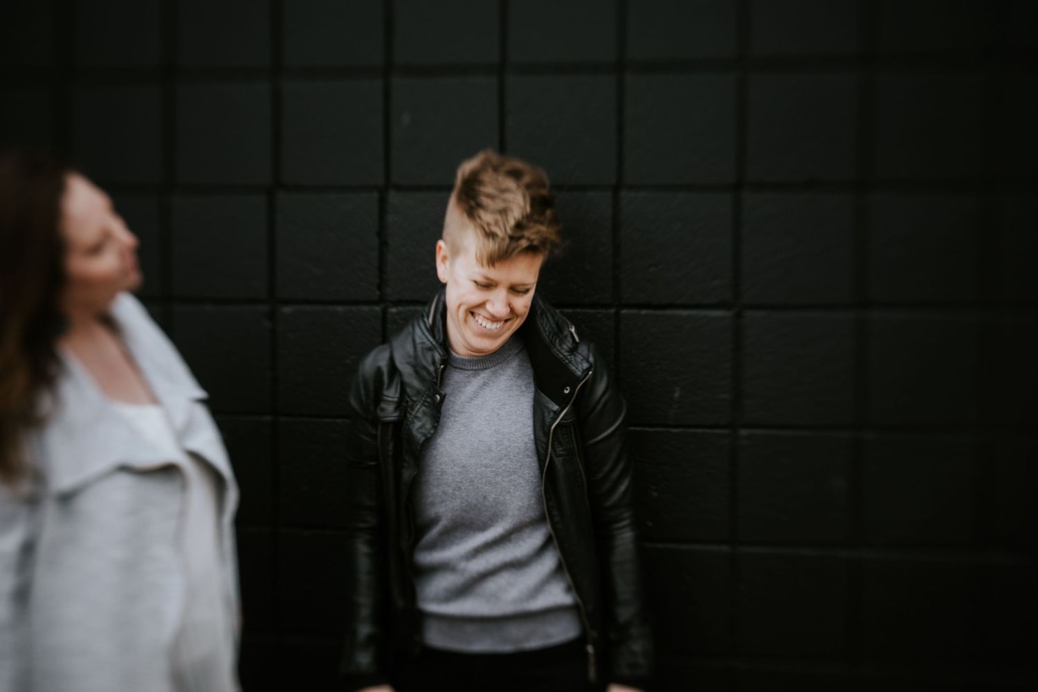 People in casual attire stand and laugh together against a dark wall in a candid moment.