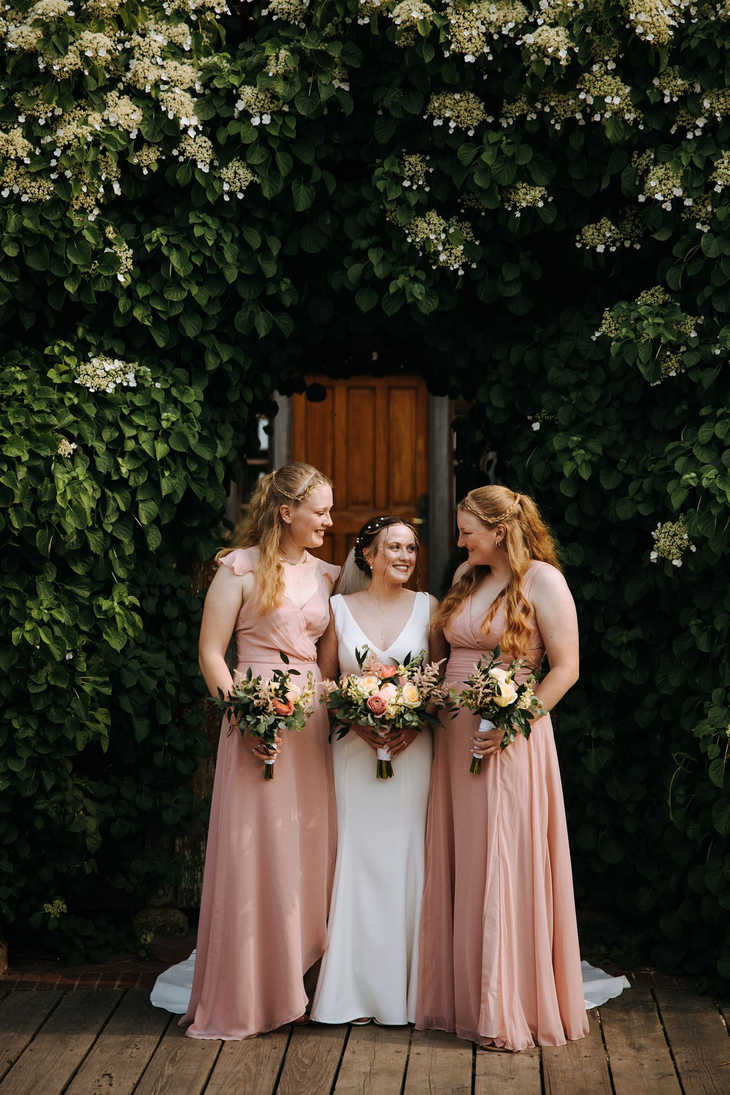 Three women in formal dresses pose with bouquets against a floral-covered wall.