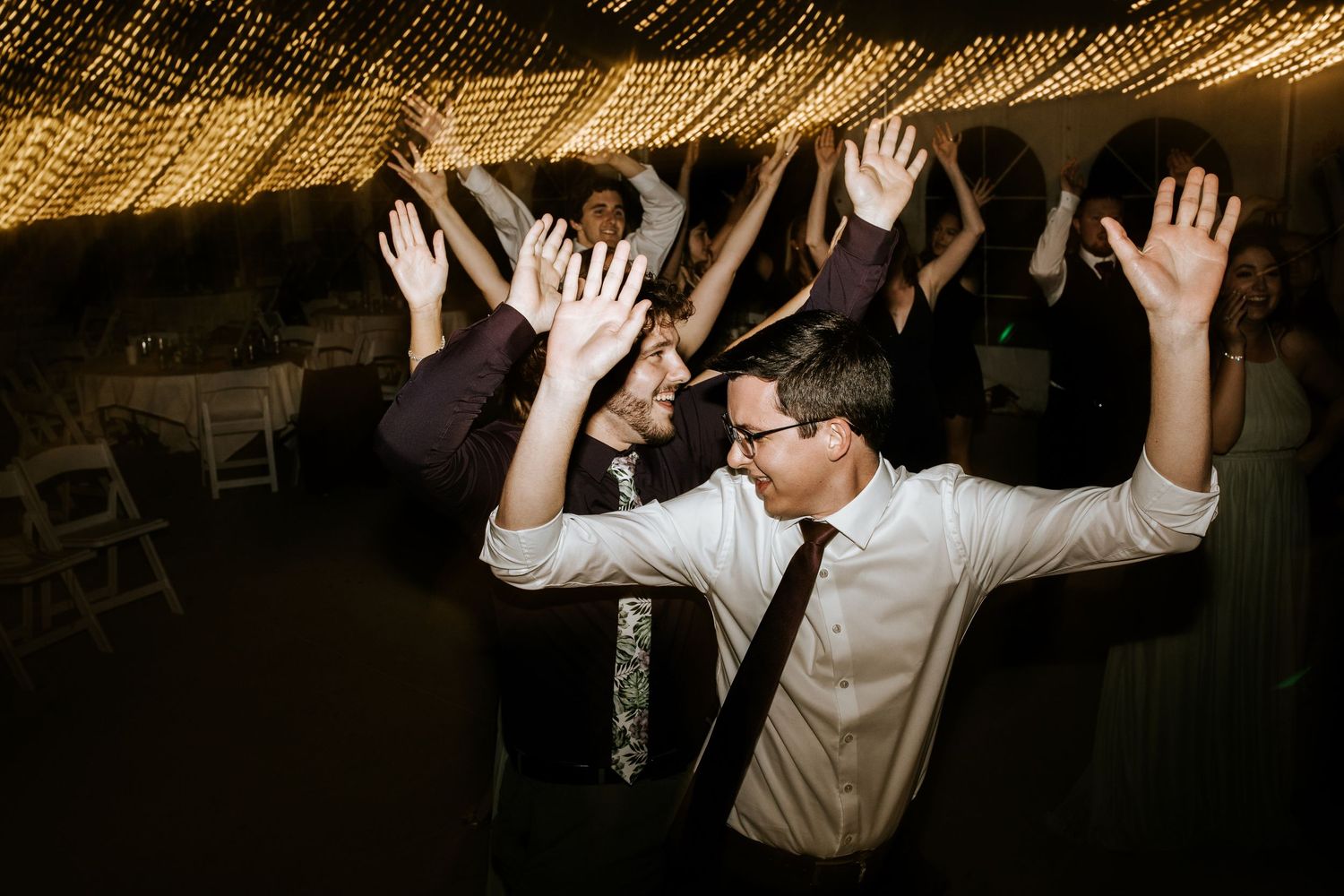 Energetic dance party with guests raising hands under warm lighting at a wedding reception.