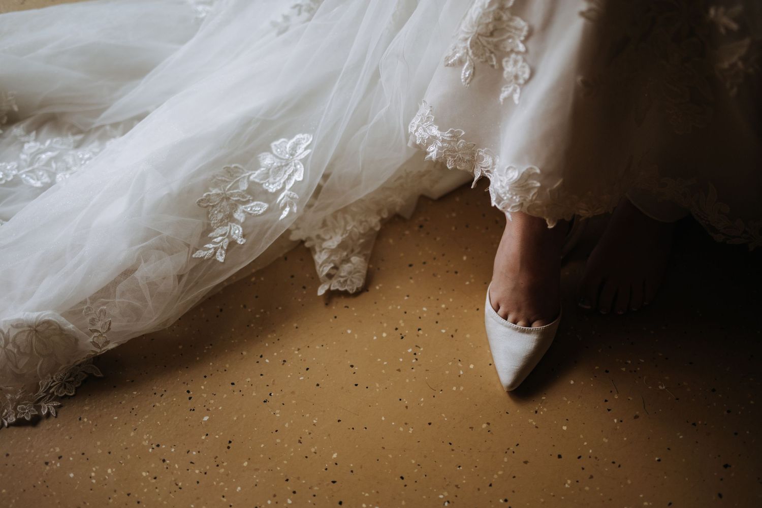 Close-up view of a flowing white wedding dress hem against wooden flooring.