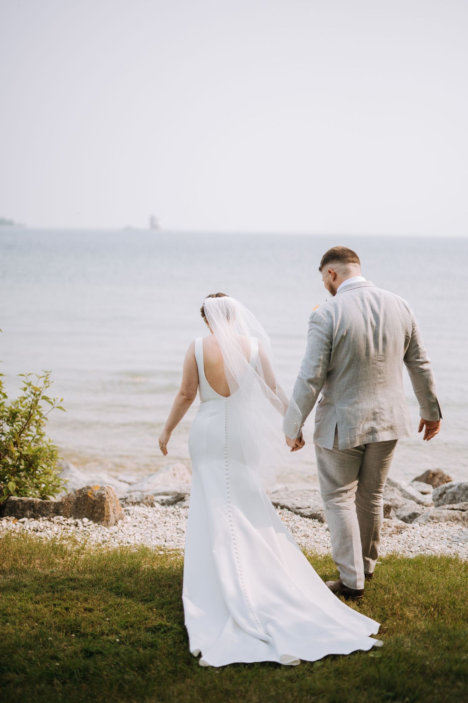 A couple in wedding attire walks along a rocky beach shoreline.