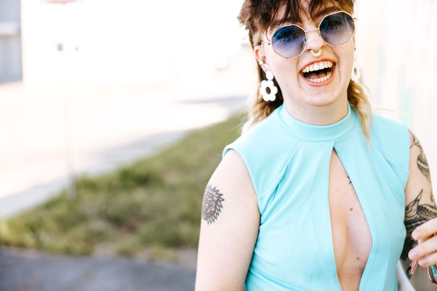 Person in turquoise sleeveless top and sunglasses laughing joyfully while showing tattoos outdoors.