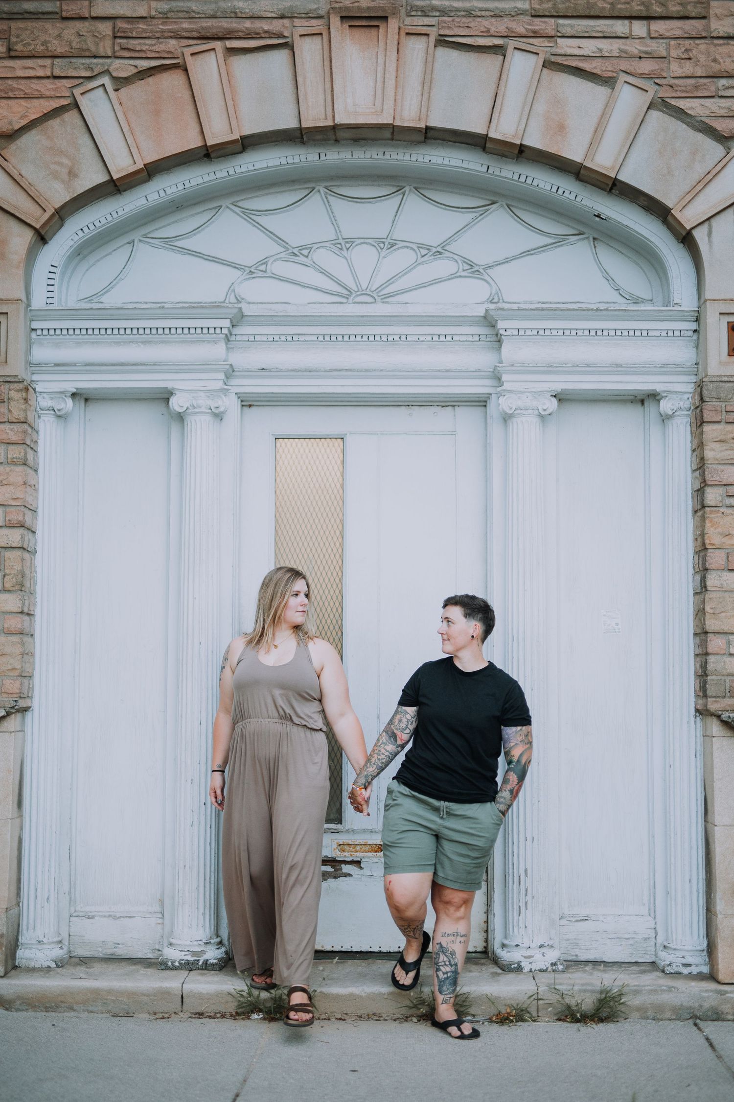 Couple poses casually against white architectural arches and stone wall.