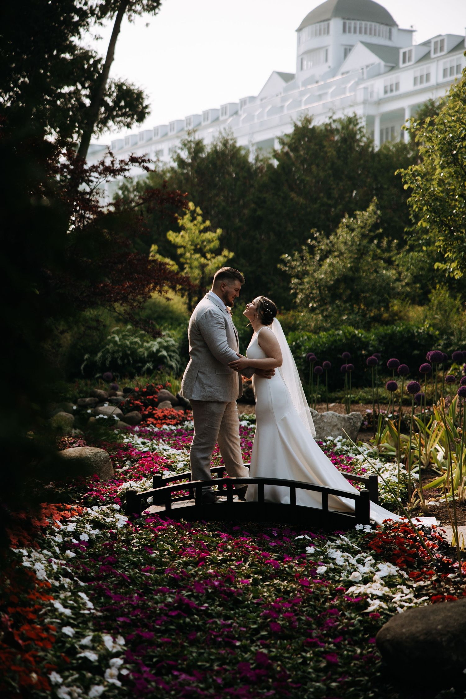 Couple poses on garden bridge surrounded by colorful flowers with majestic white hotel in background.