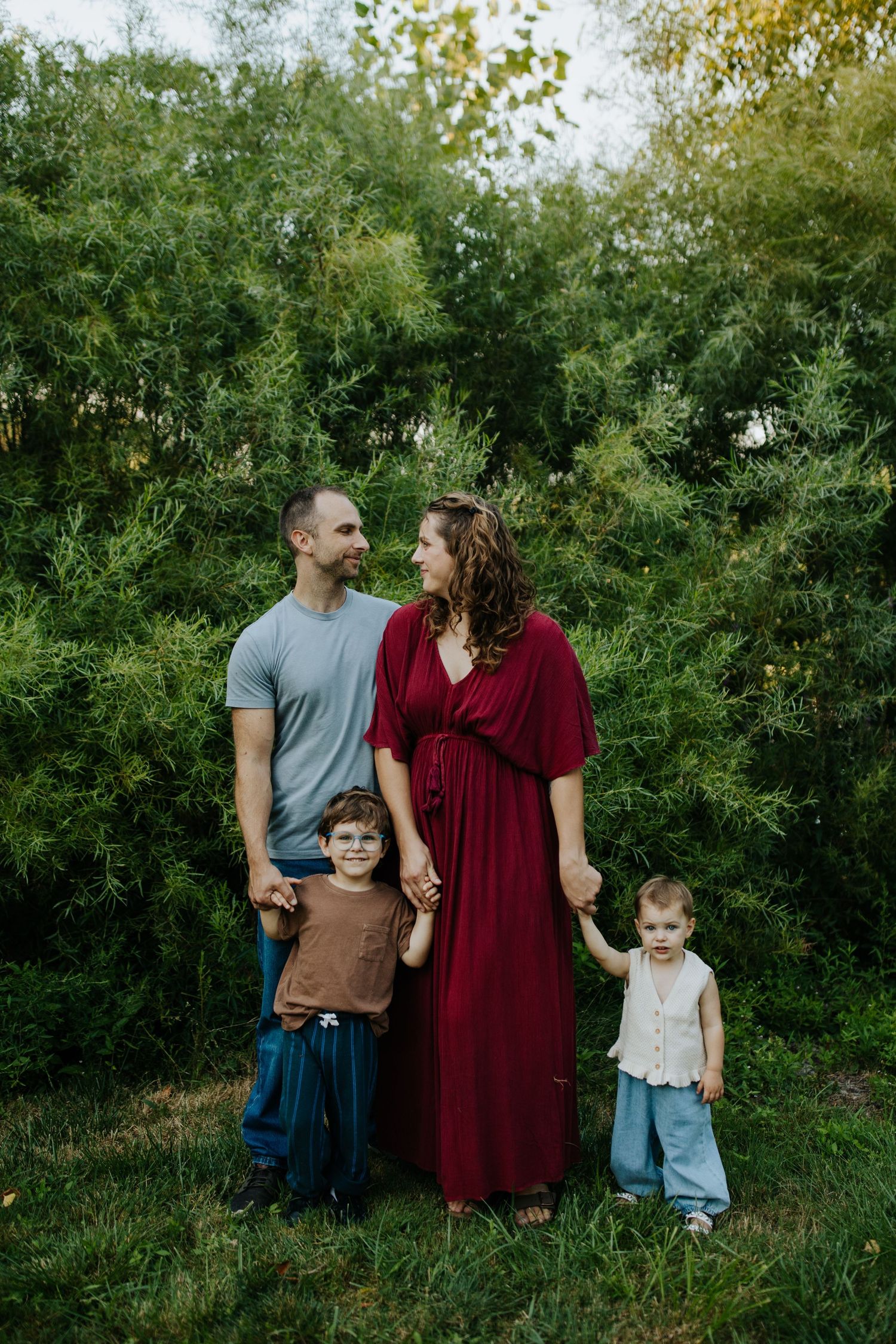 A couple and child walk together through lush greenery wearing burgundy and casual attire.