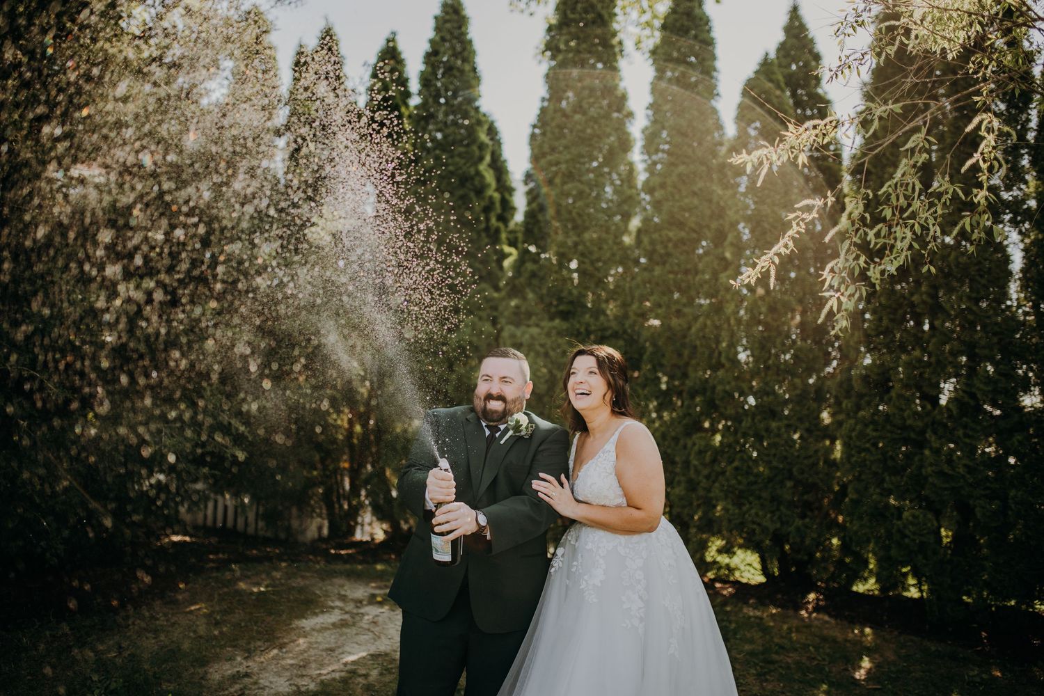 A wedding couple celebrates with champagne spray in a garden setting at sunset.